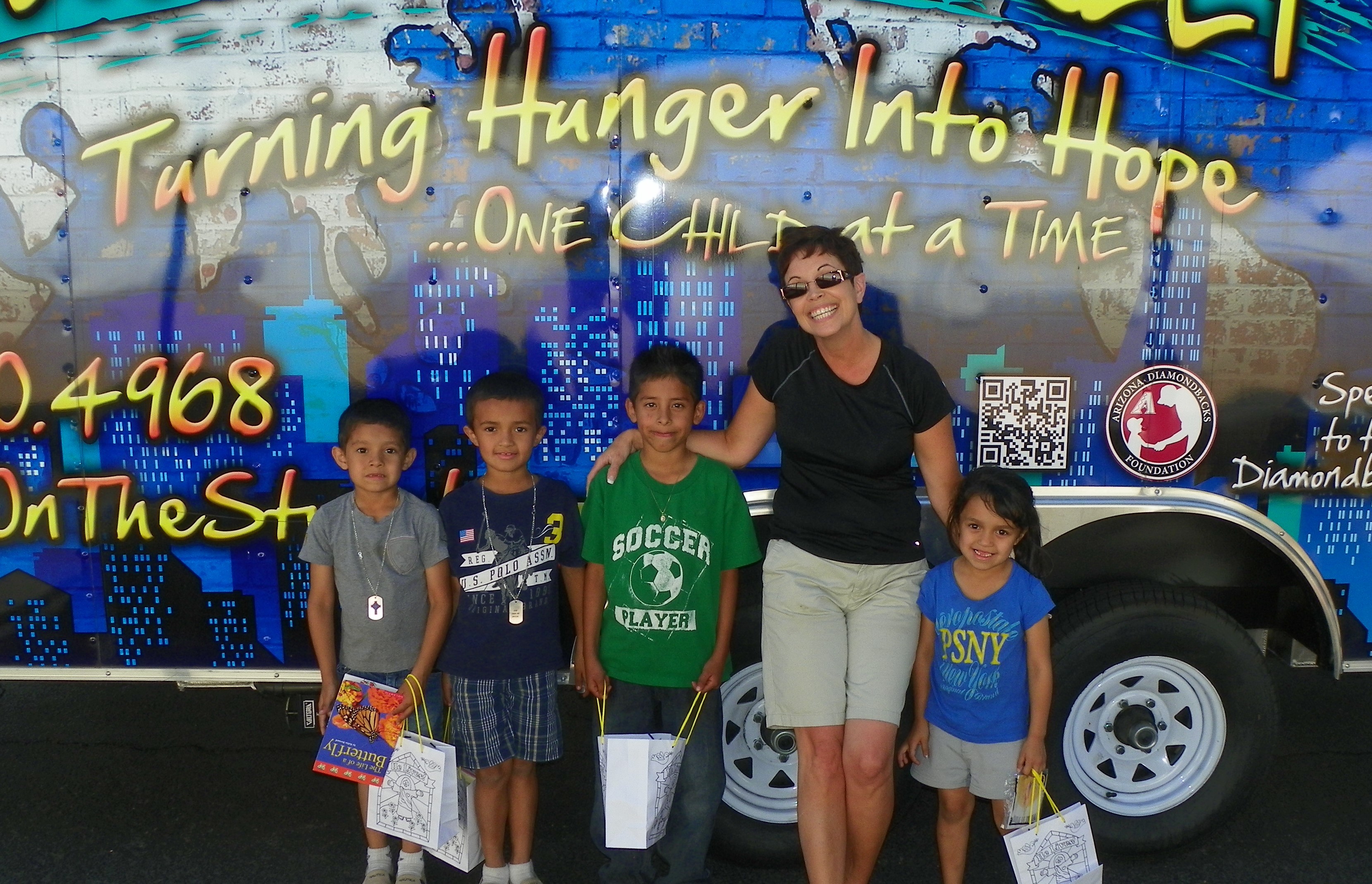 Four children and an adult woman stand in front of a colorful bus with text and graphics, holding papers or bags.