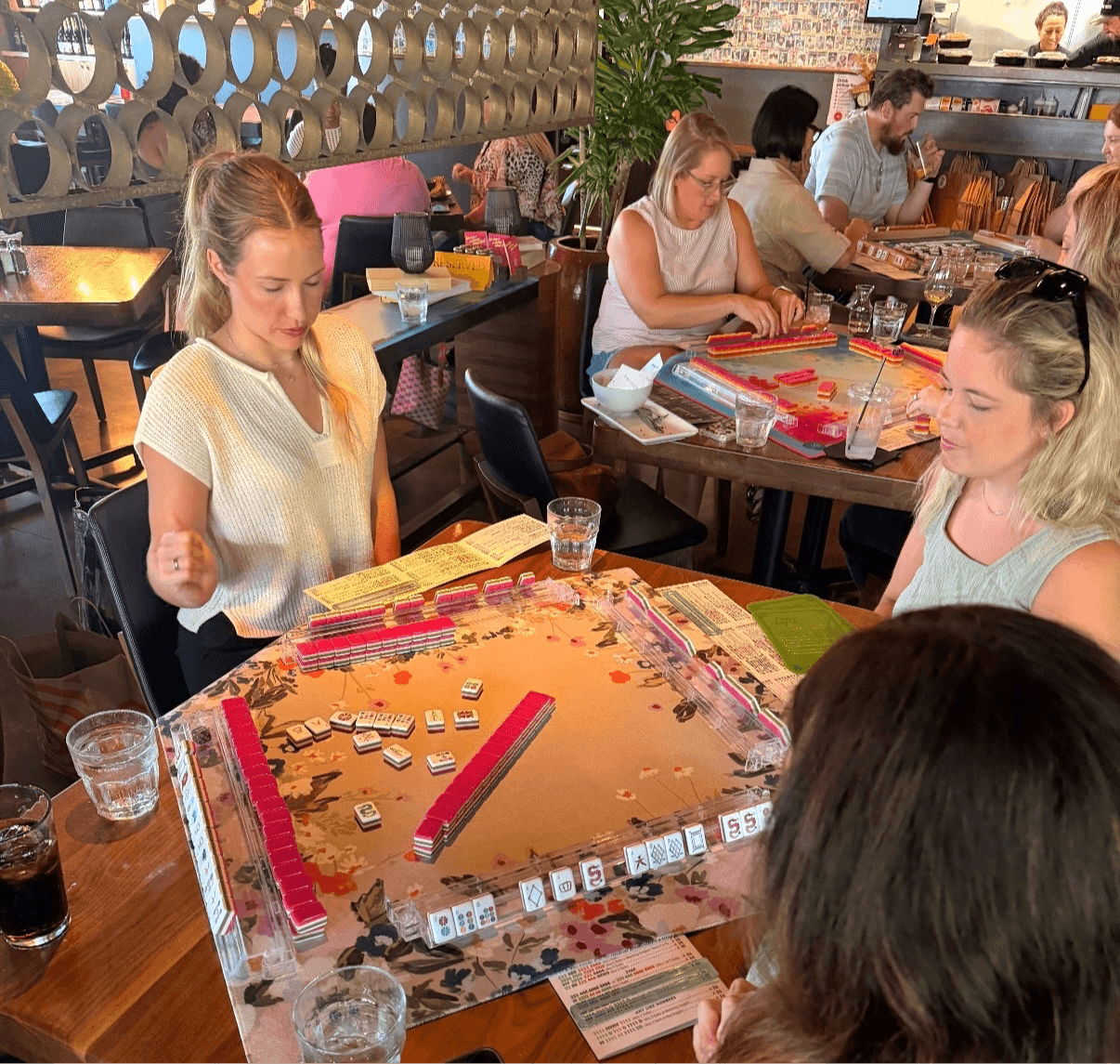 Four women sit around a table playing a board game in a lively restaurant or cafe, with other patrons in the background.