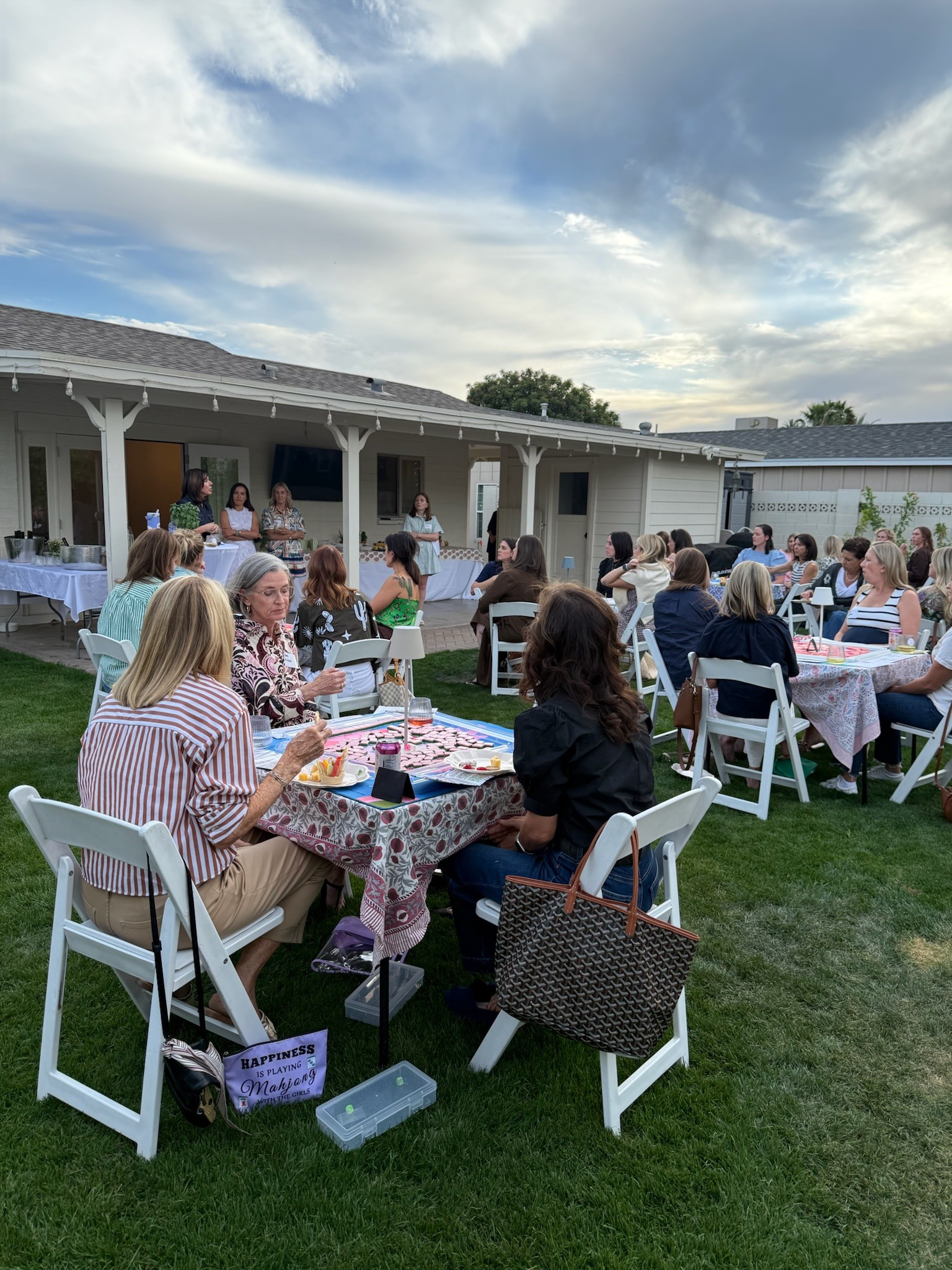 People seated at tables outdoors during daytime, with a building and sky in the background.