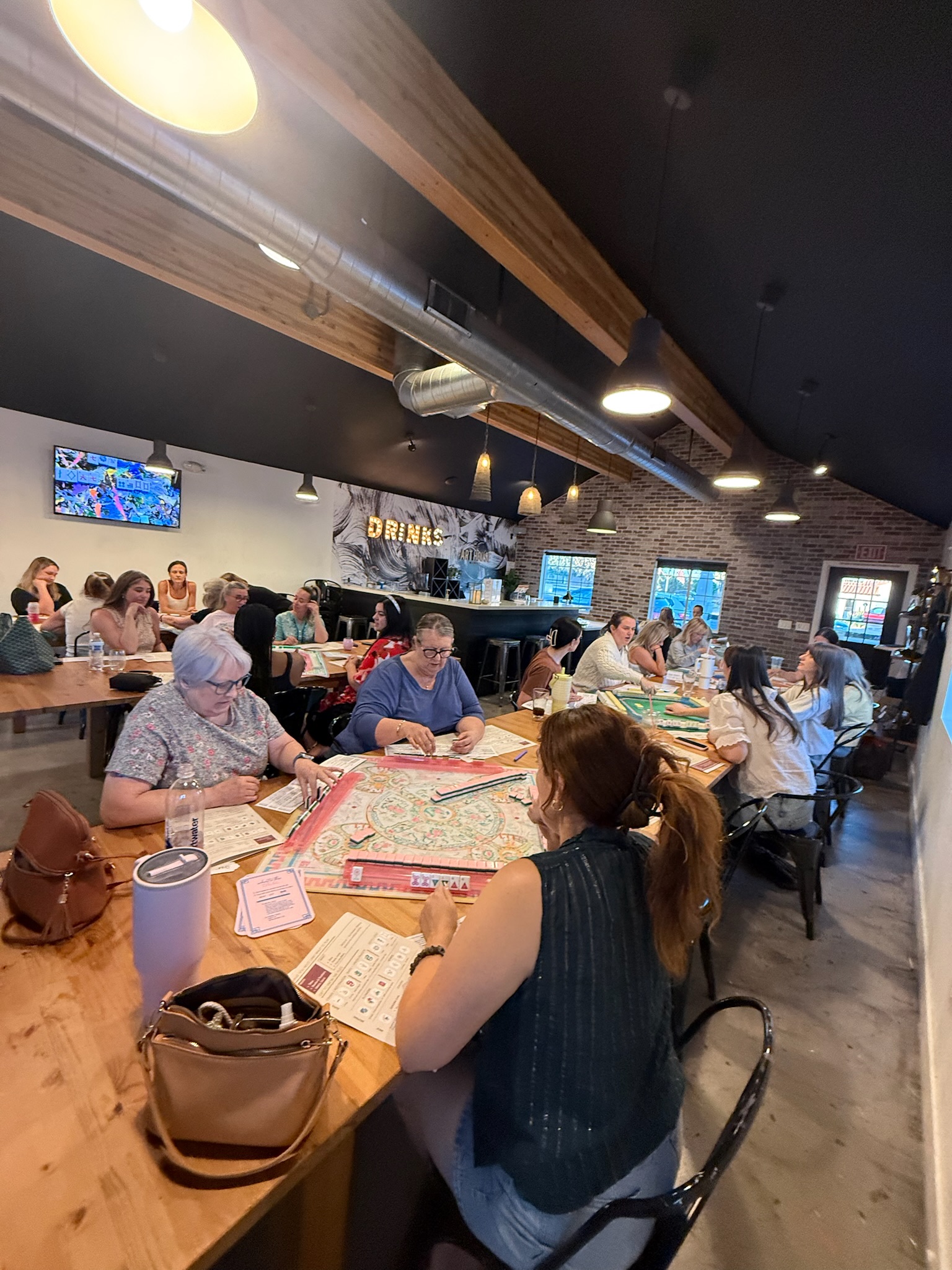 People sitting at tables in a restaurant, some wearing masks, with TVs on the wall and a brick wall in the background.