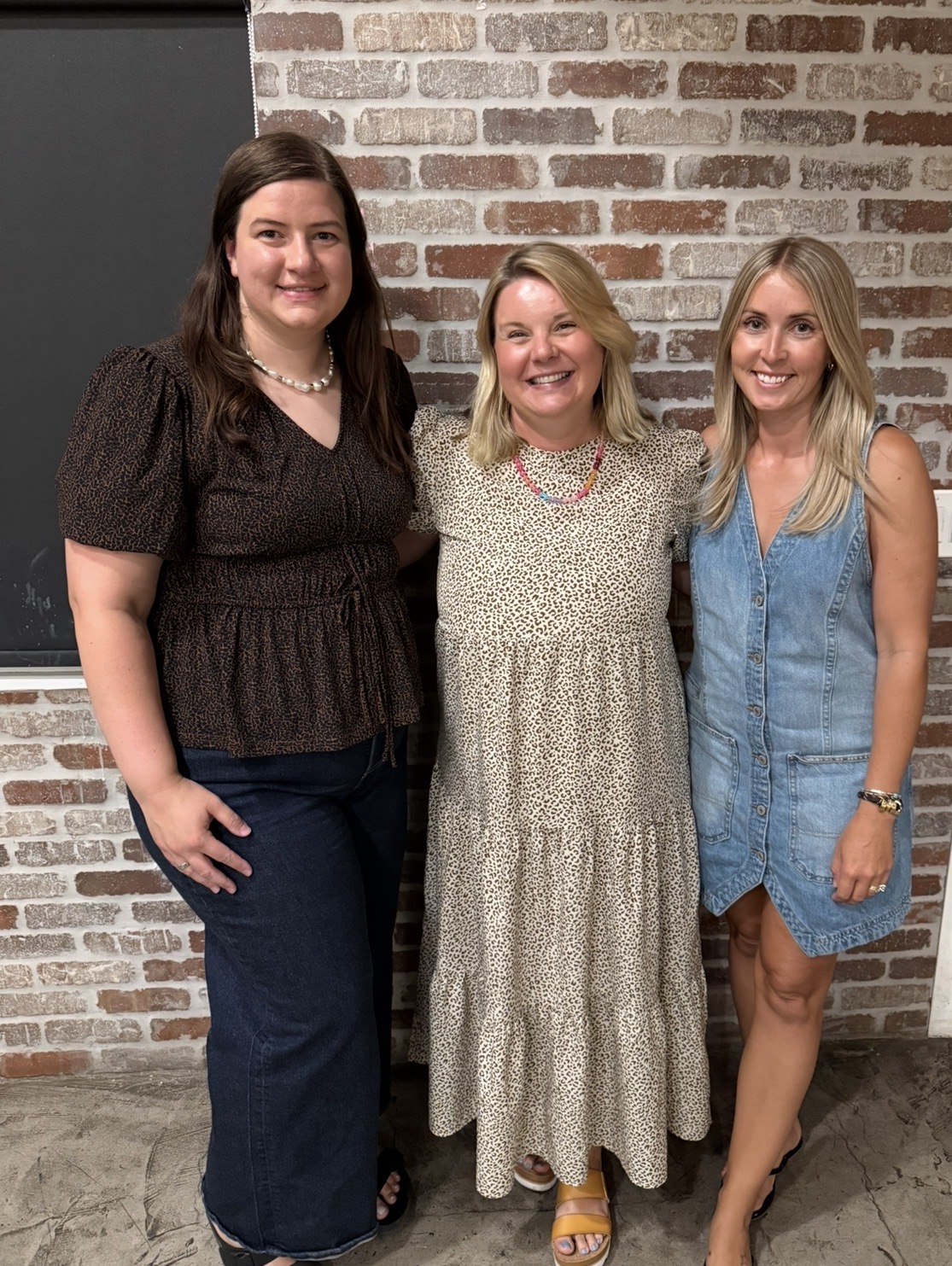 Three women standing together in front of a brick wall, smiling, with casual clothing. Word count: 20