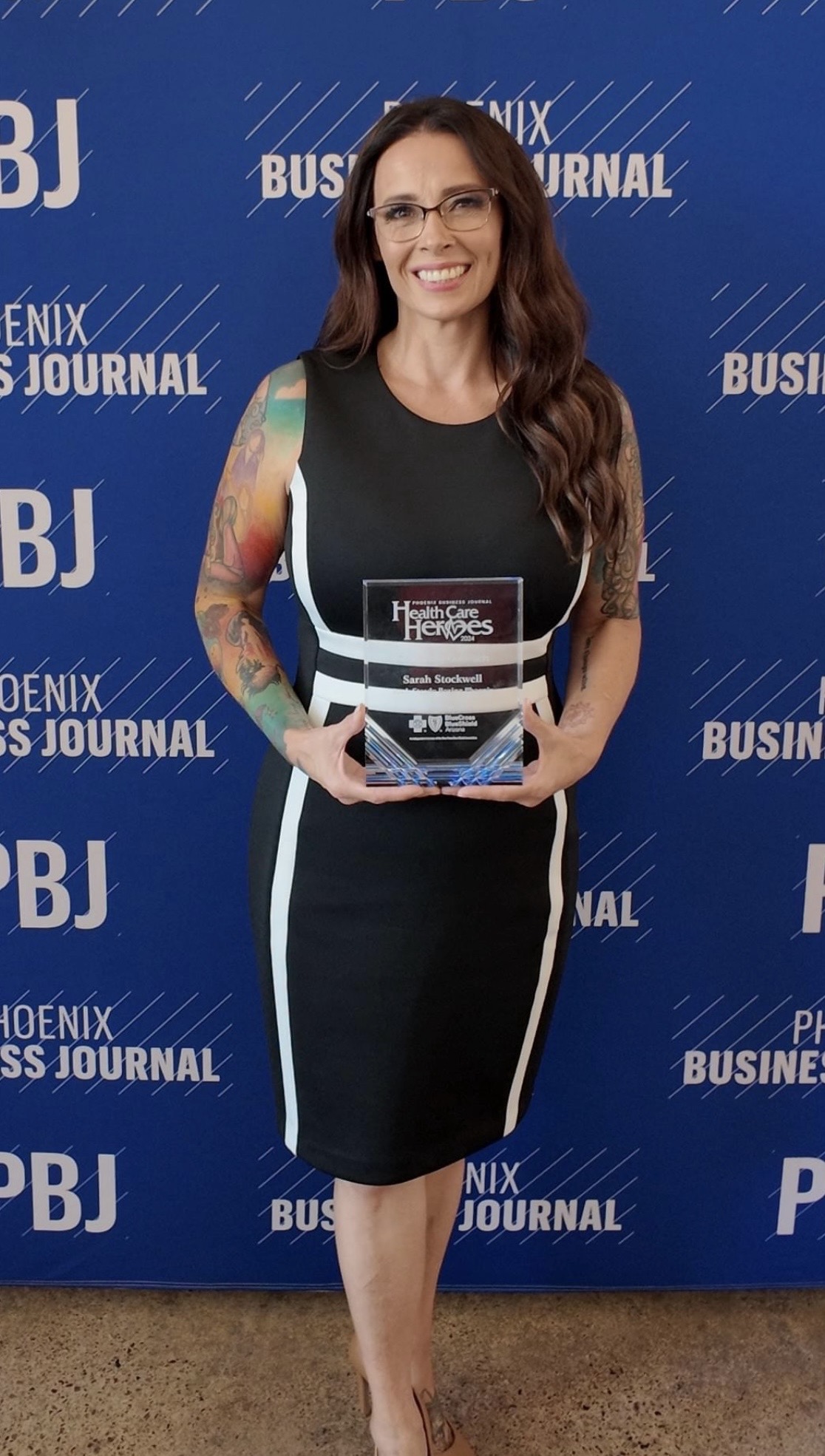 Woman with long hair and glasses holding an award, standing in front of a blue backdrop with text, smiling.