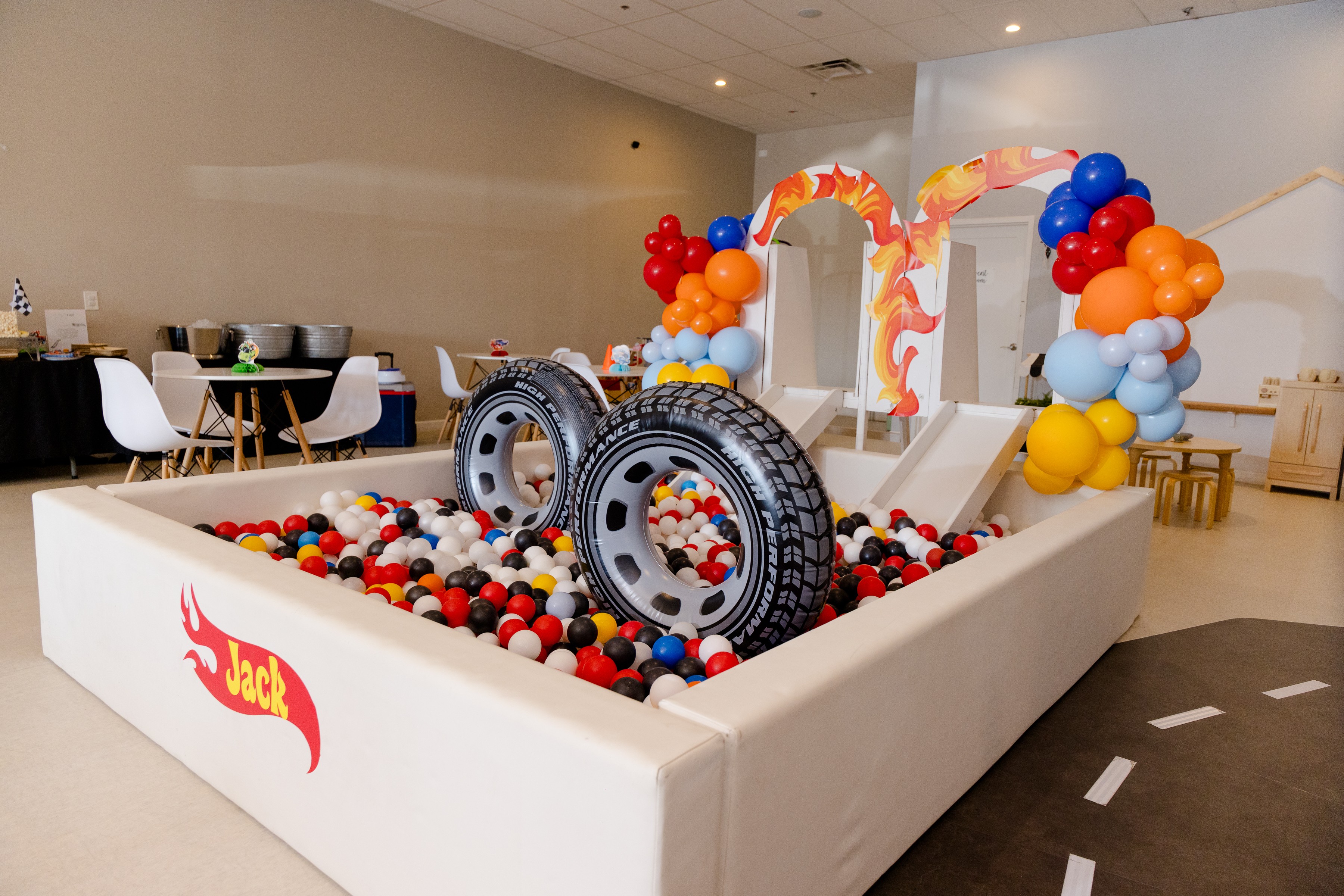 Indoor party setup with large tires, colorful balloons, and a slide, decorated for children, with tables and chairs in background.