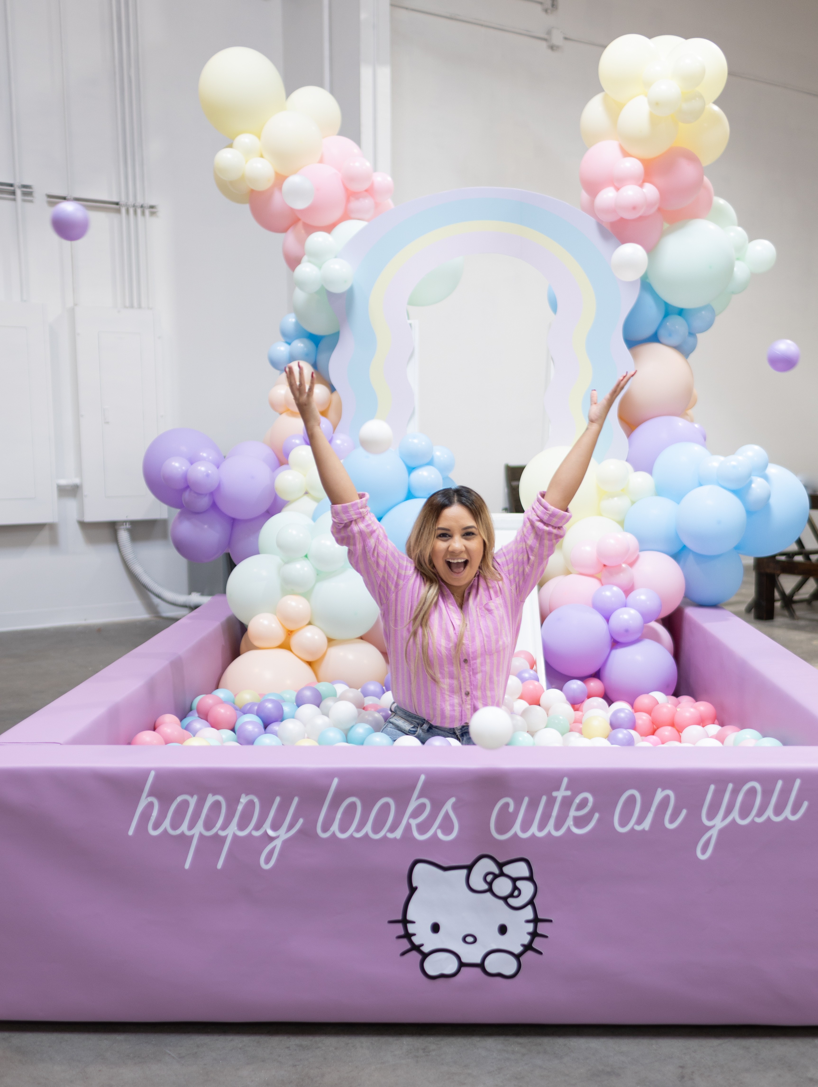 Child smiling with arms raised in a colorful balloon-filled play area with a Hello Kitty design, inside a pink enclosure.