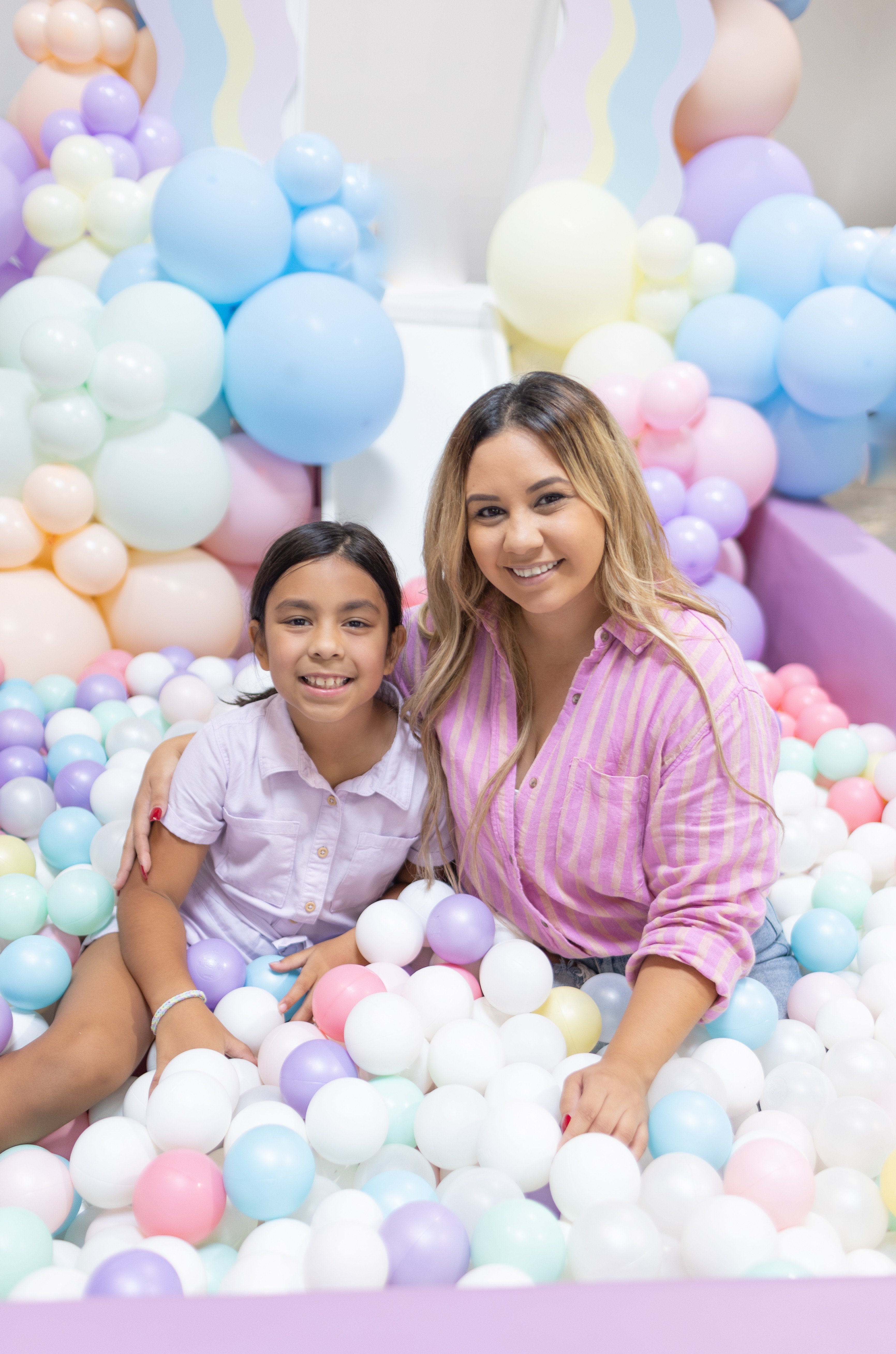 Two smiling females, one young girl and one woman, sitting among pastel-colored balloons in a decorated setting.