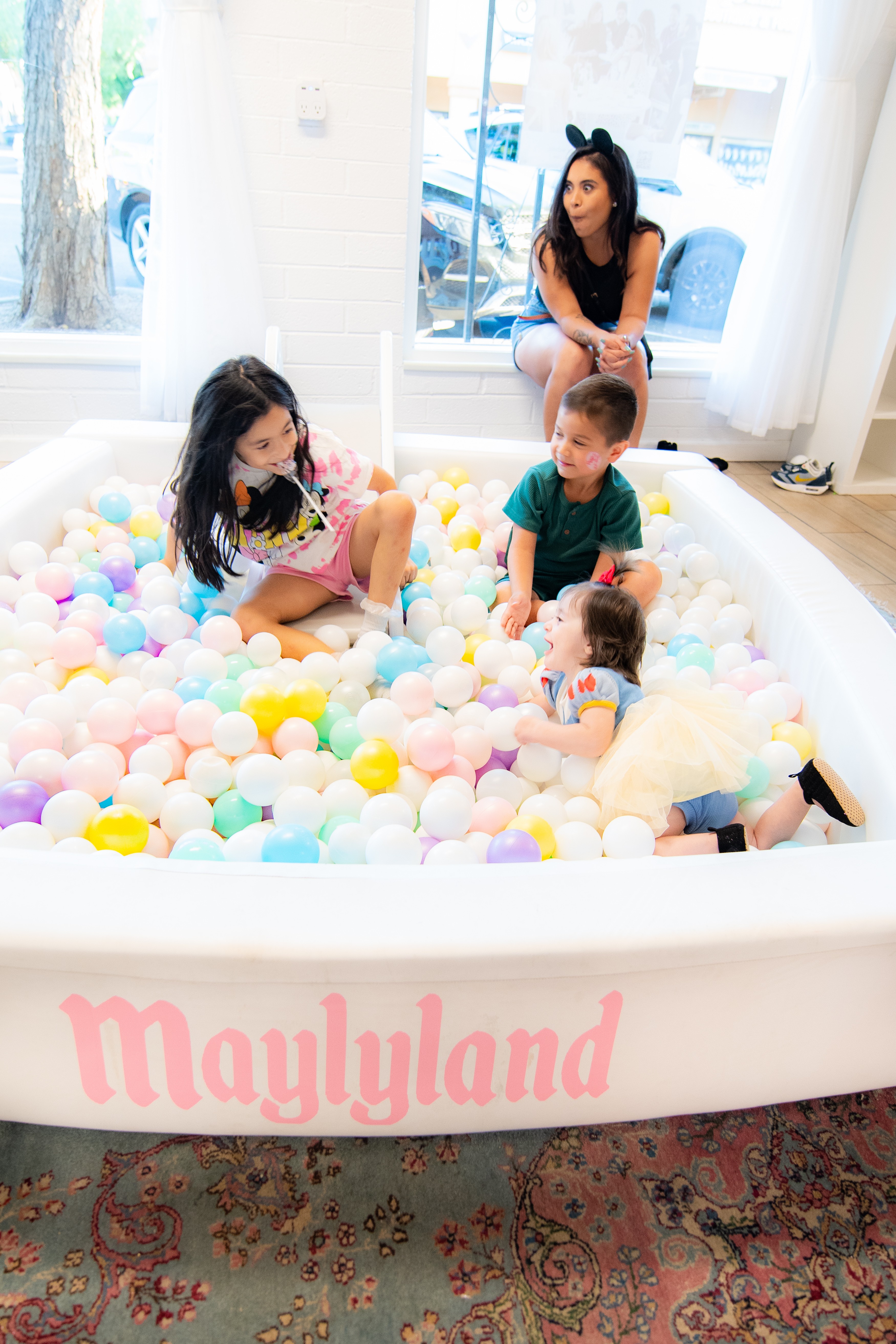 Four children and an adult in a ball pit with colorful balls, inside a bright room with large windows, Maylyland written on the side.