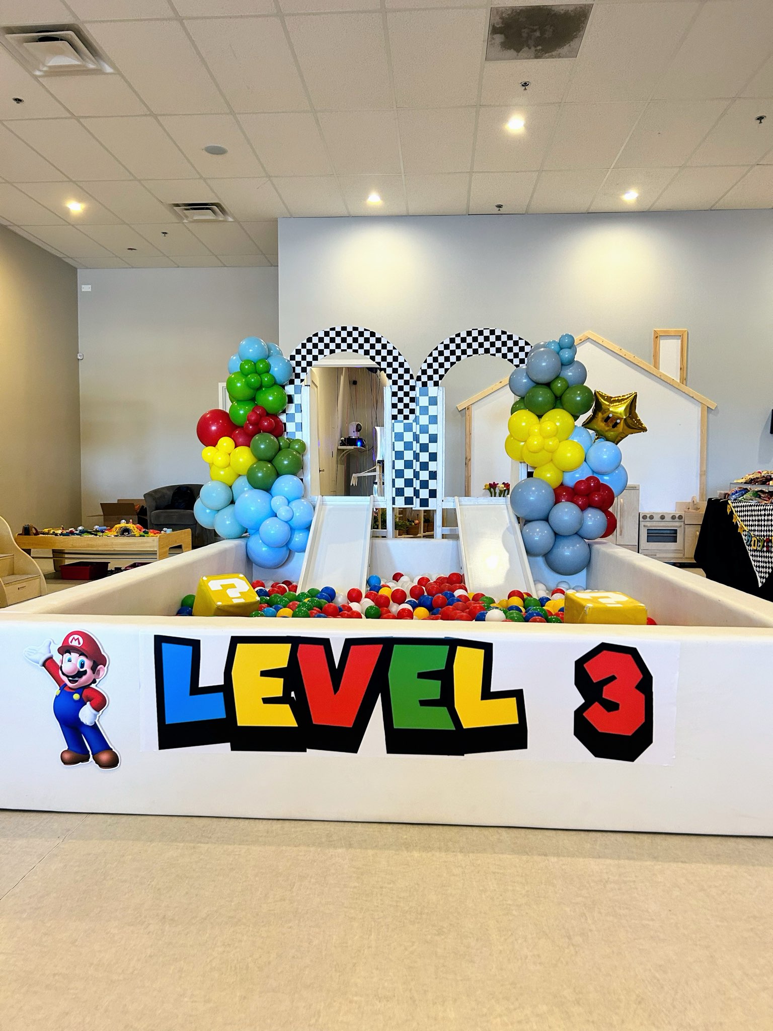 Party setup with colorful balloon decorations, a sign reading 'LEVEL 3' with a Mario character, and a backdrop with checkered arches.