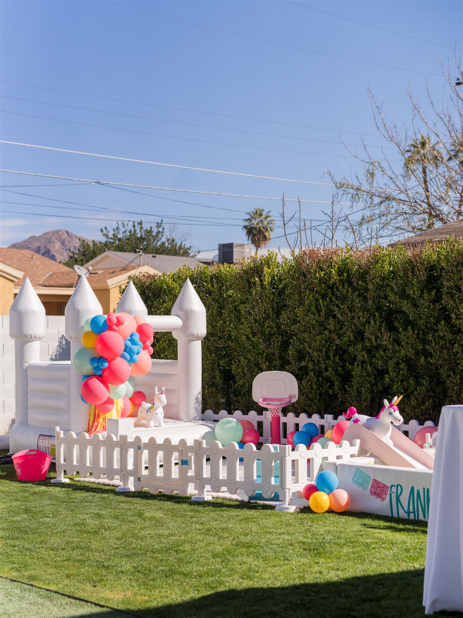 Colorful balloons decorate a white picket fence in a backyard with green grass and a hedge, under a clear blue sky.