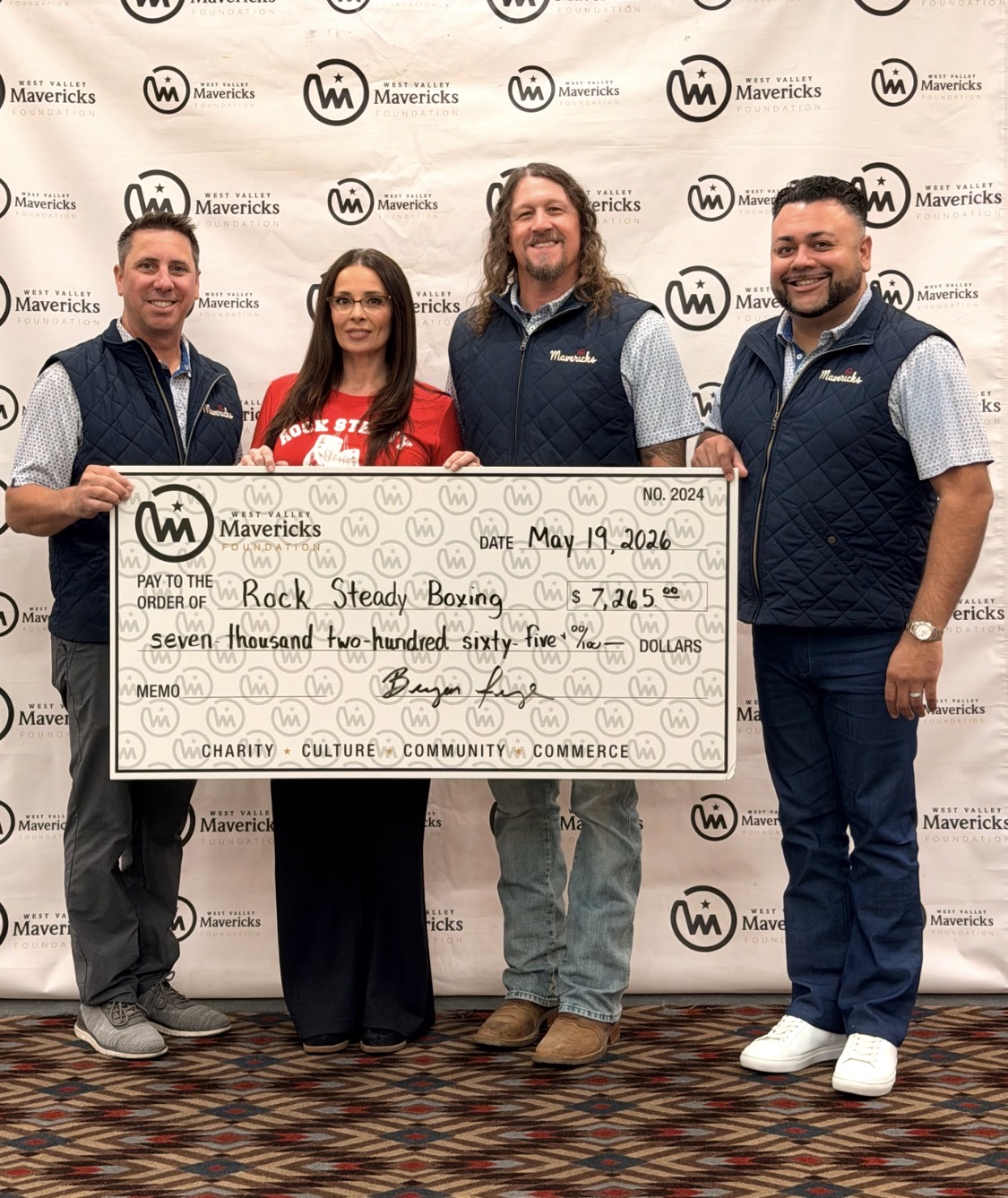 Four people holding a large check in front of a backdrop with logos, smiling, on a patterned carpeted floor.
