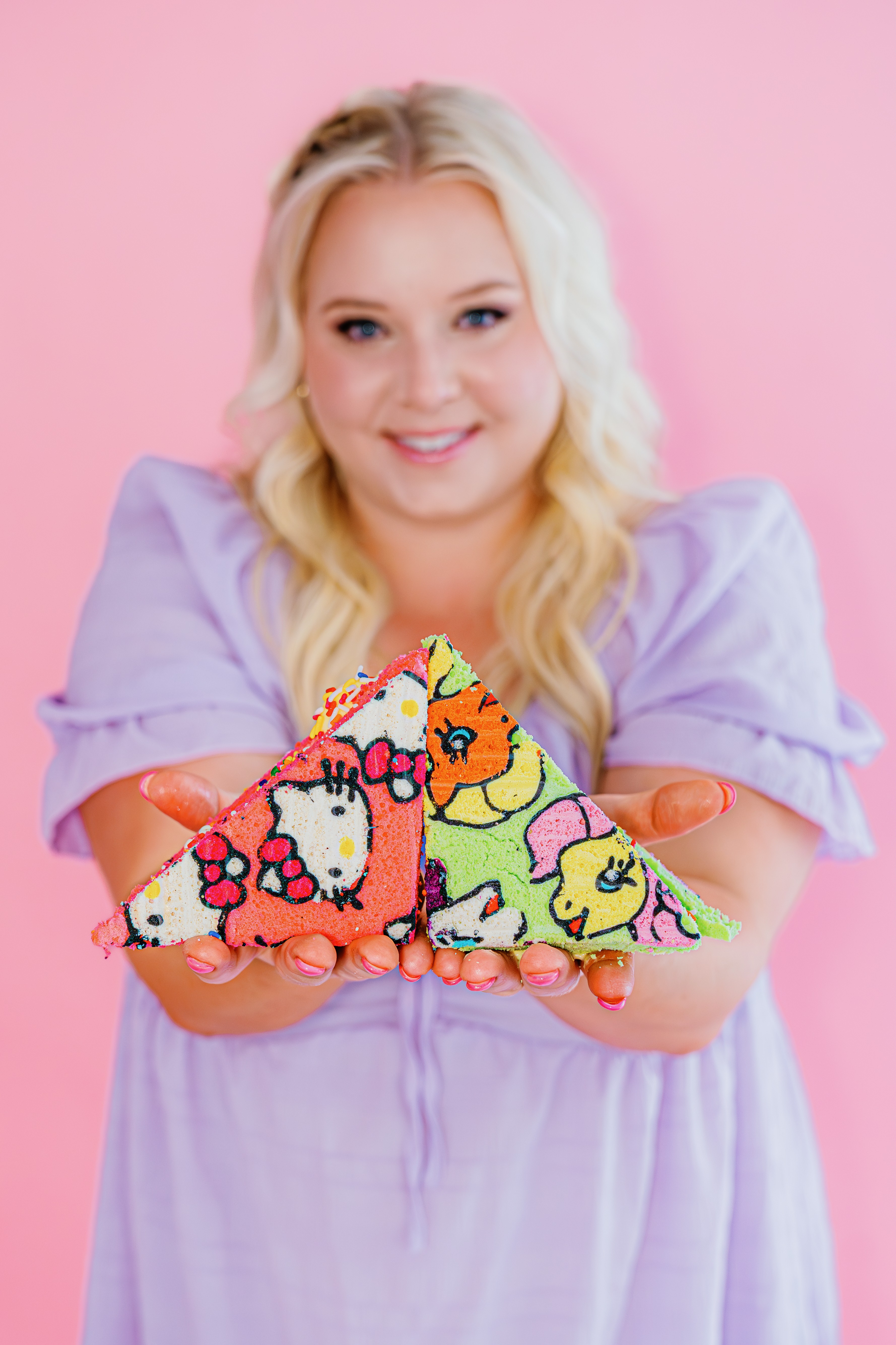Young woman with blonde hair holding colorful unicorn plush toys, wearing a lavender top, against a pink background.