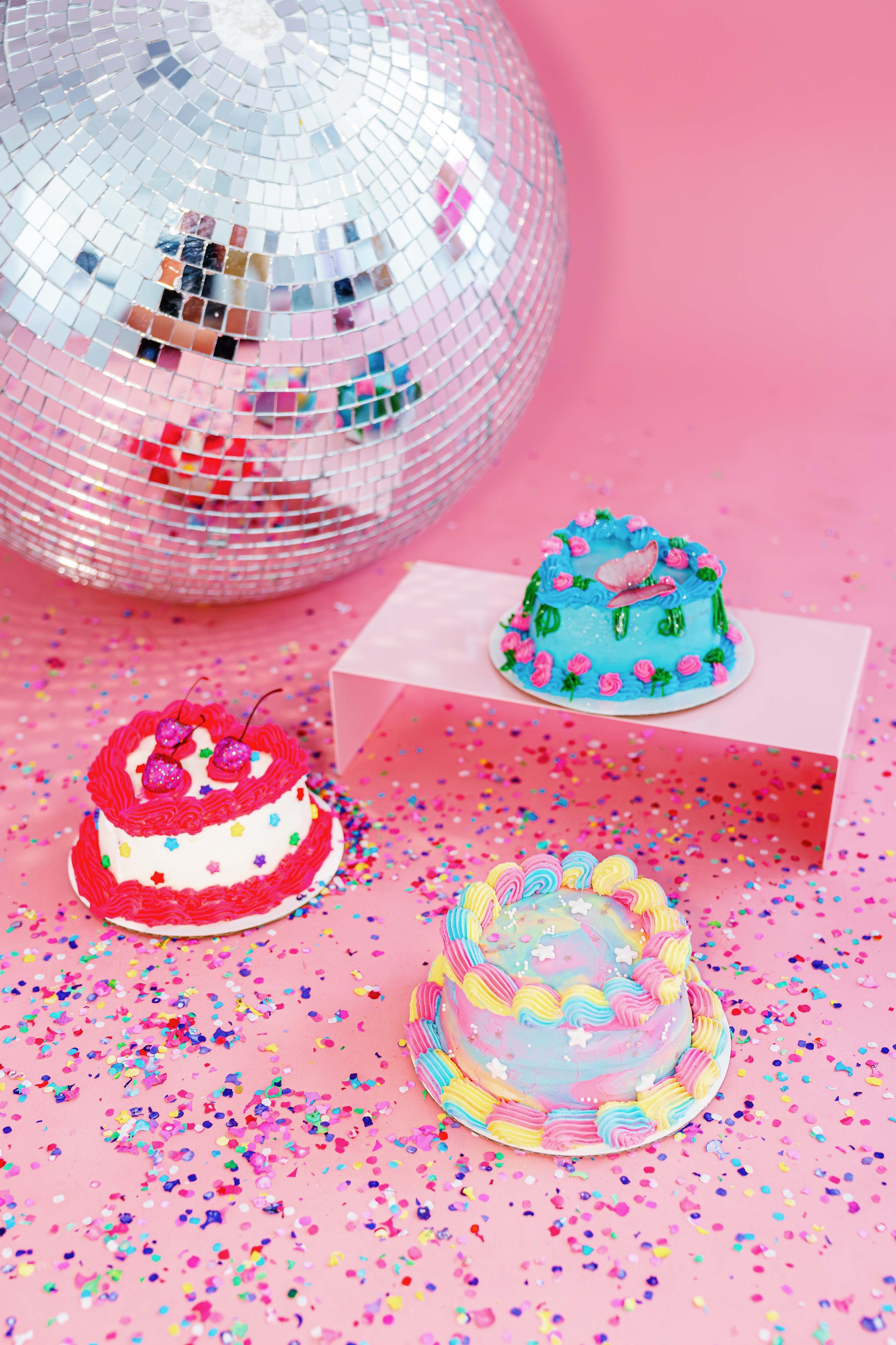 Three small decorated cakes on a pink surface with colorful confetti, a disco ball in the background, and a pink box.