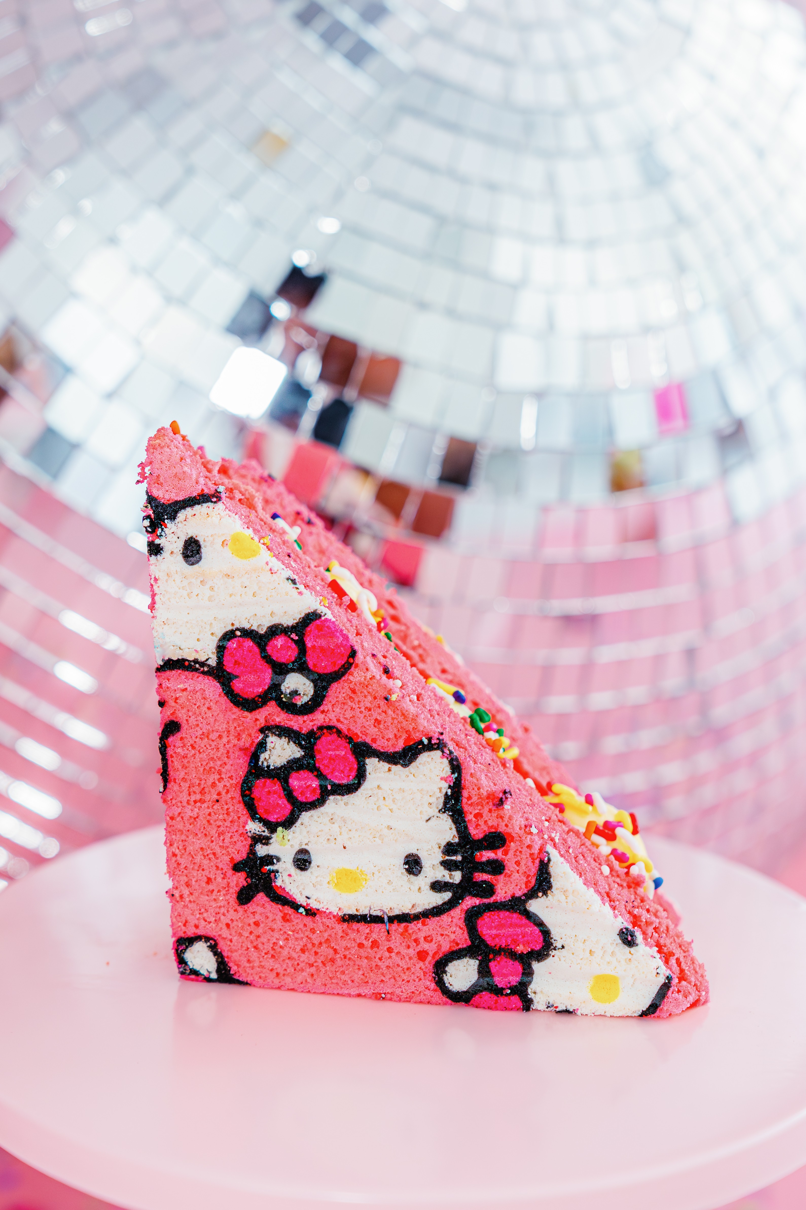 Decorative cake slice with Hello Kitty design, pink and white colors, on a pink plate, with a reflective disco ball background.