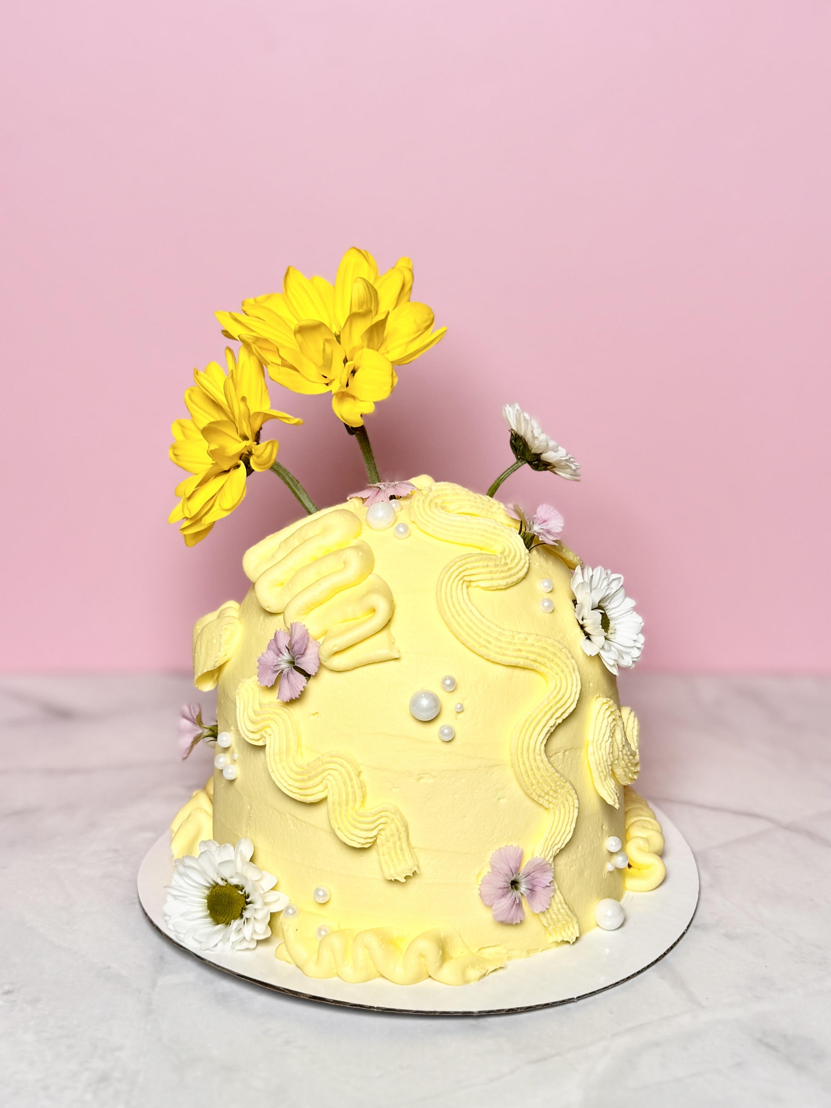 Yellow decorated cake with flowers on a marble surface against pink background.