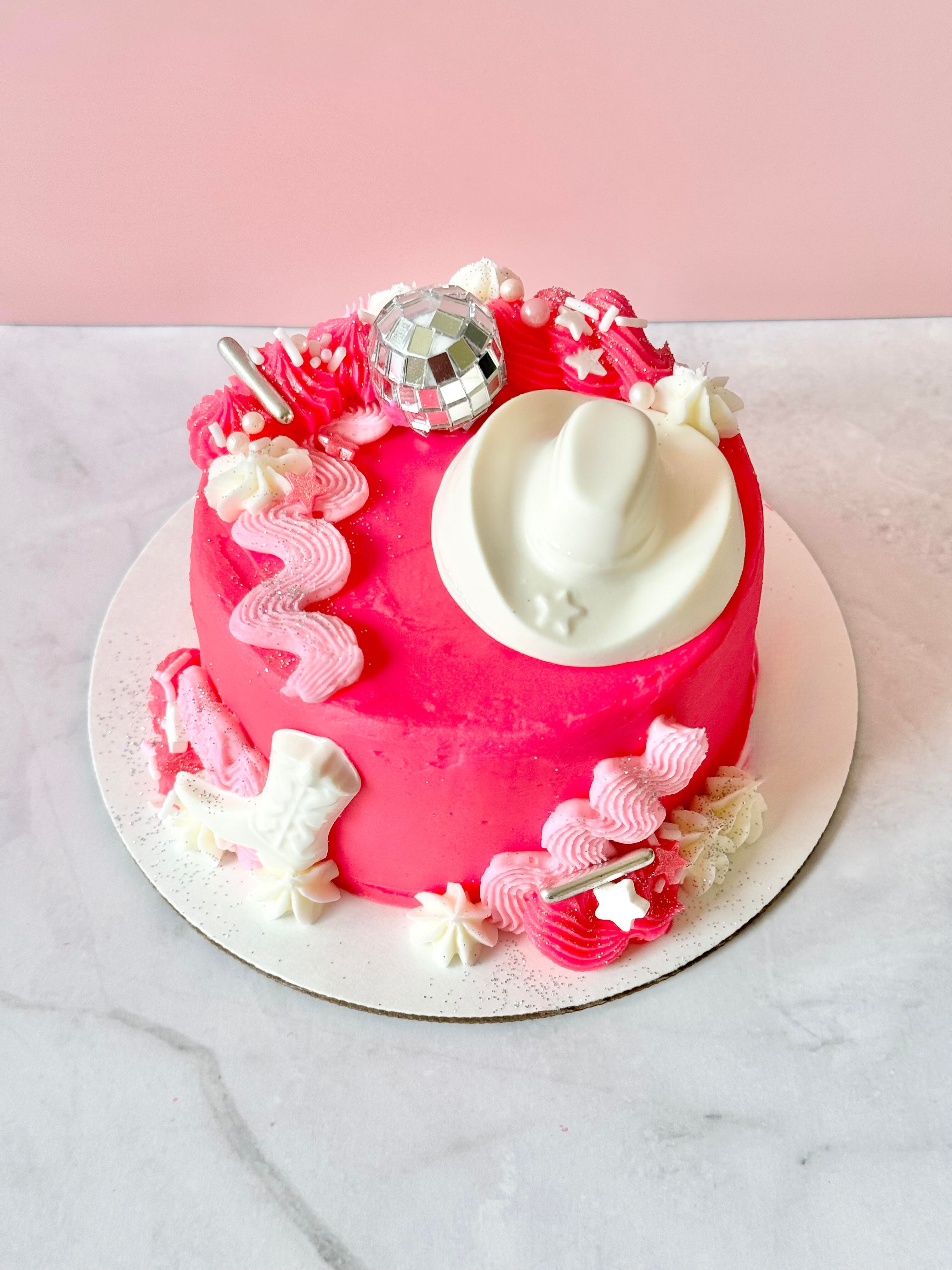 Round pink cake decorated with white and pink frosting, a white hat, and shiny silver and pink ornaments, on a white surface.