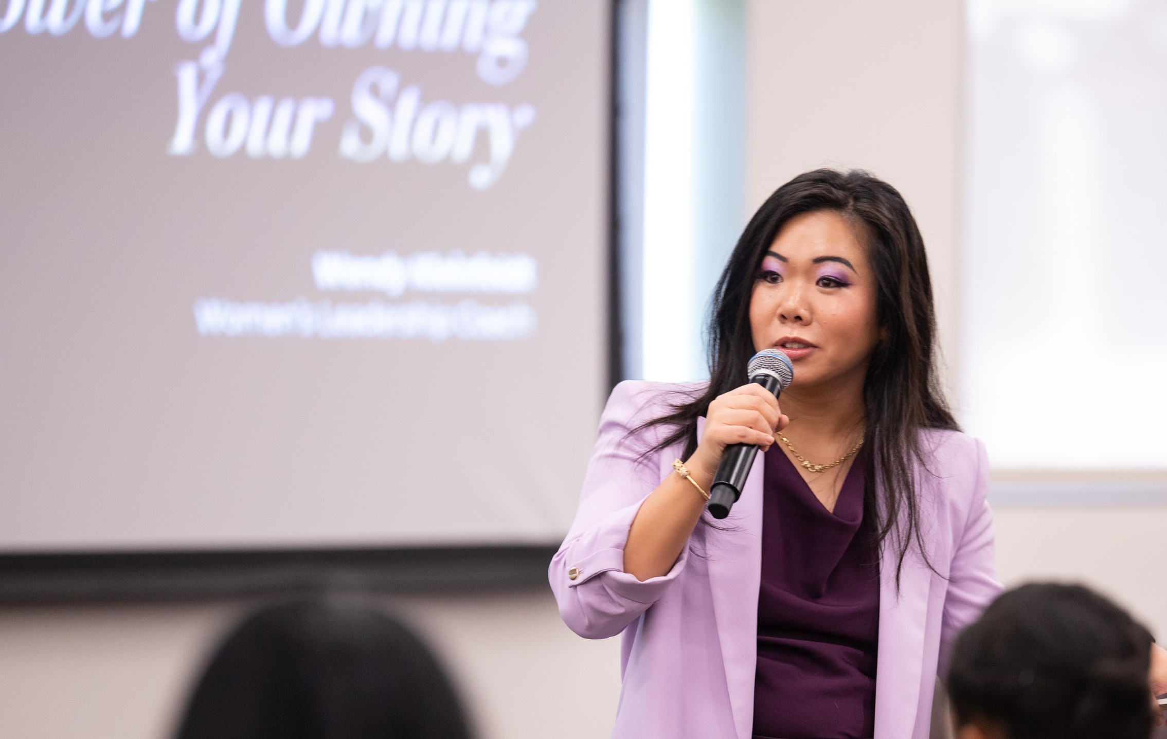 Woman speaking into a microphone during a presentation, with a screen behind her displaying text.