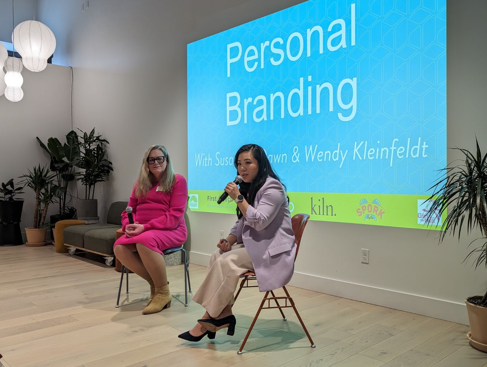 Two women seated on chairs in front of a presentation screen, one speaking into a microphone, in a room with plants and lighting.