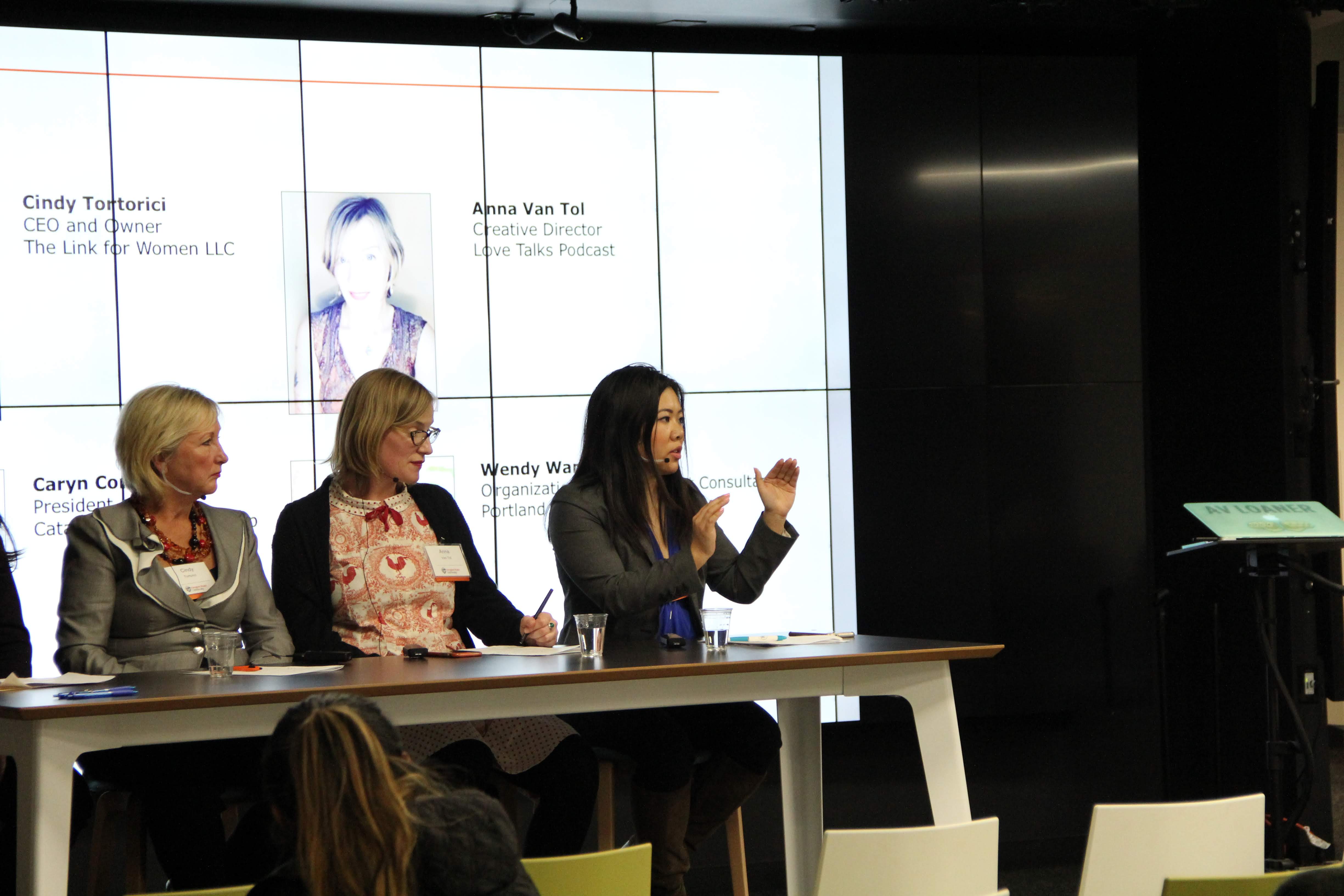 Three women seated at a panel table in front of a large screen displaying text and images, with an audience in front.