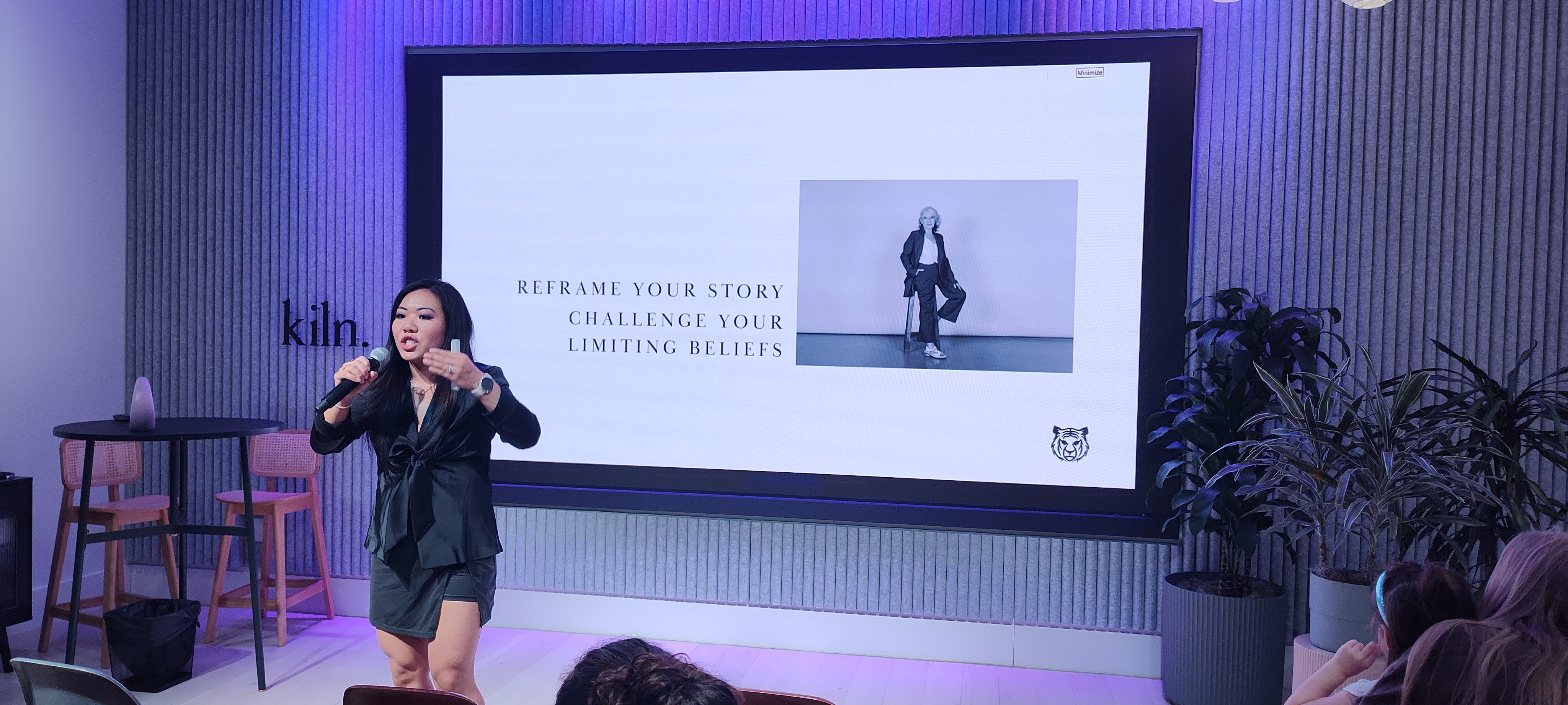 Woman speaking on stage with a large screen behind her displaying a person in a pose, audience visible in foreground.