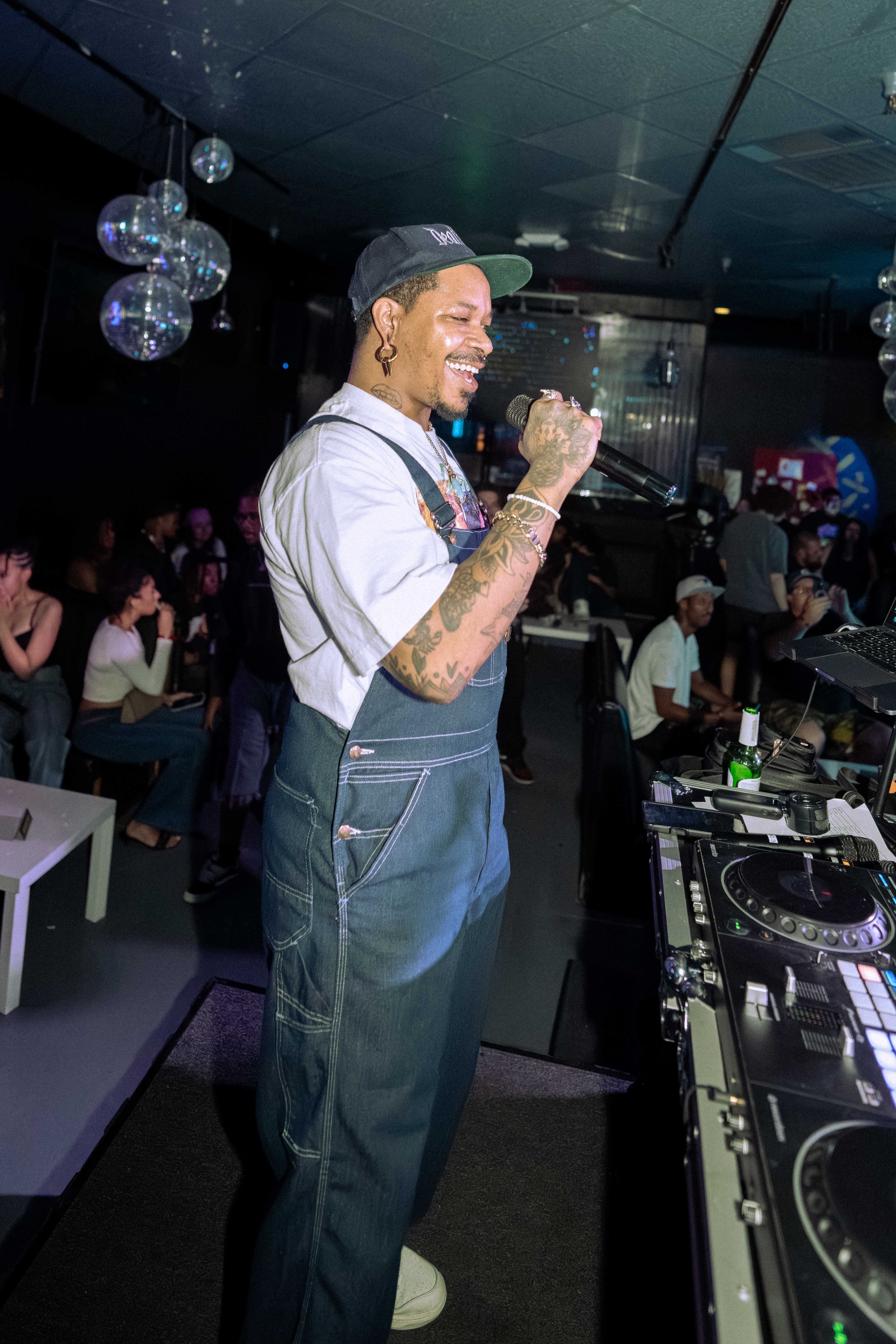 Person with tattoos and earrings, wearing a cap and overalls, smiling and holding a microphone in a club setting.