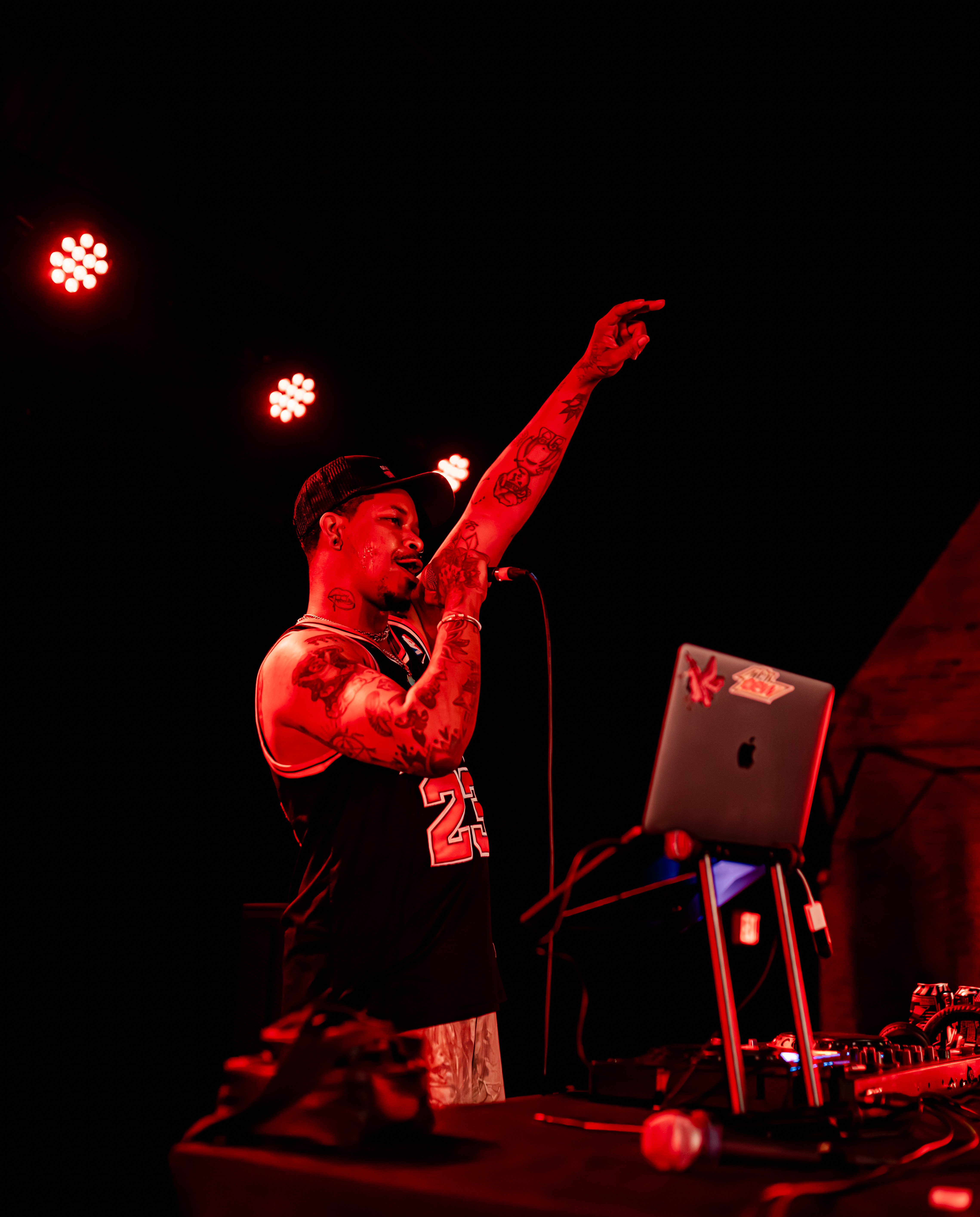 Person with tattoos and a headband performs on stage with a microphone, laptop, and DJ equipment, raising one arm.
