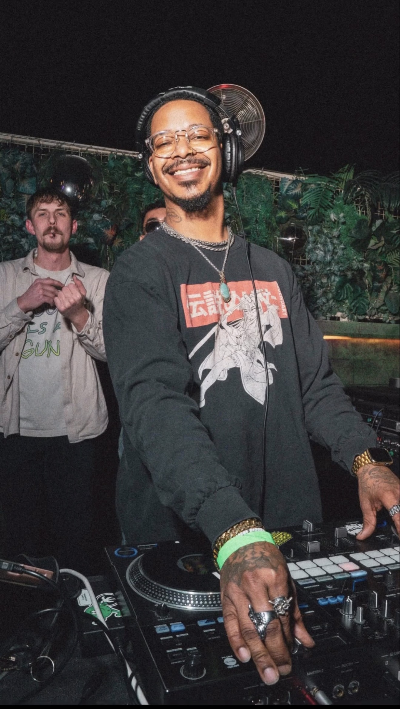 DJ smiling at turntables with a person in the background, night scene, outdoor setting, colorful lights, casual clothing