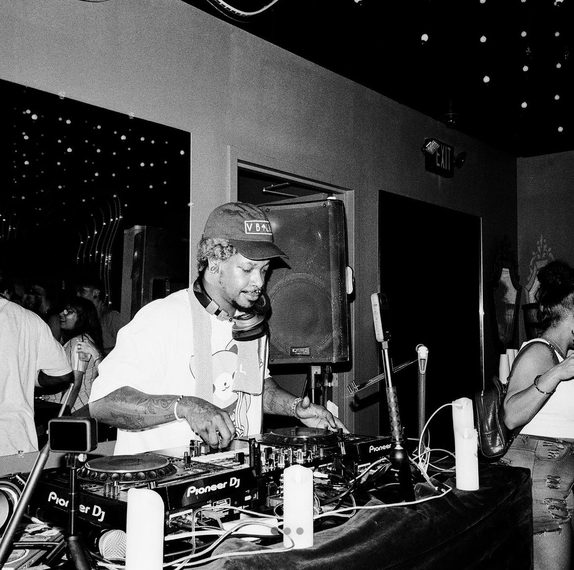 DJ with headphones operating turntables and equipment at a party or event, with people in the background, in a dimly lit room.