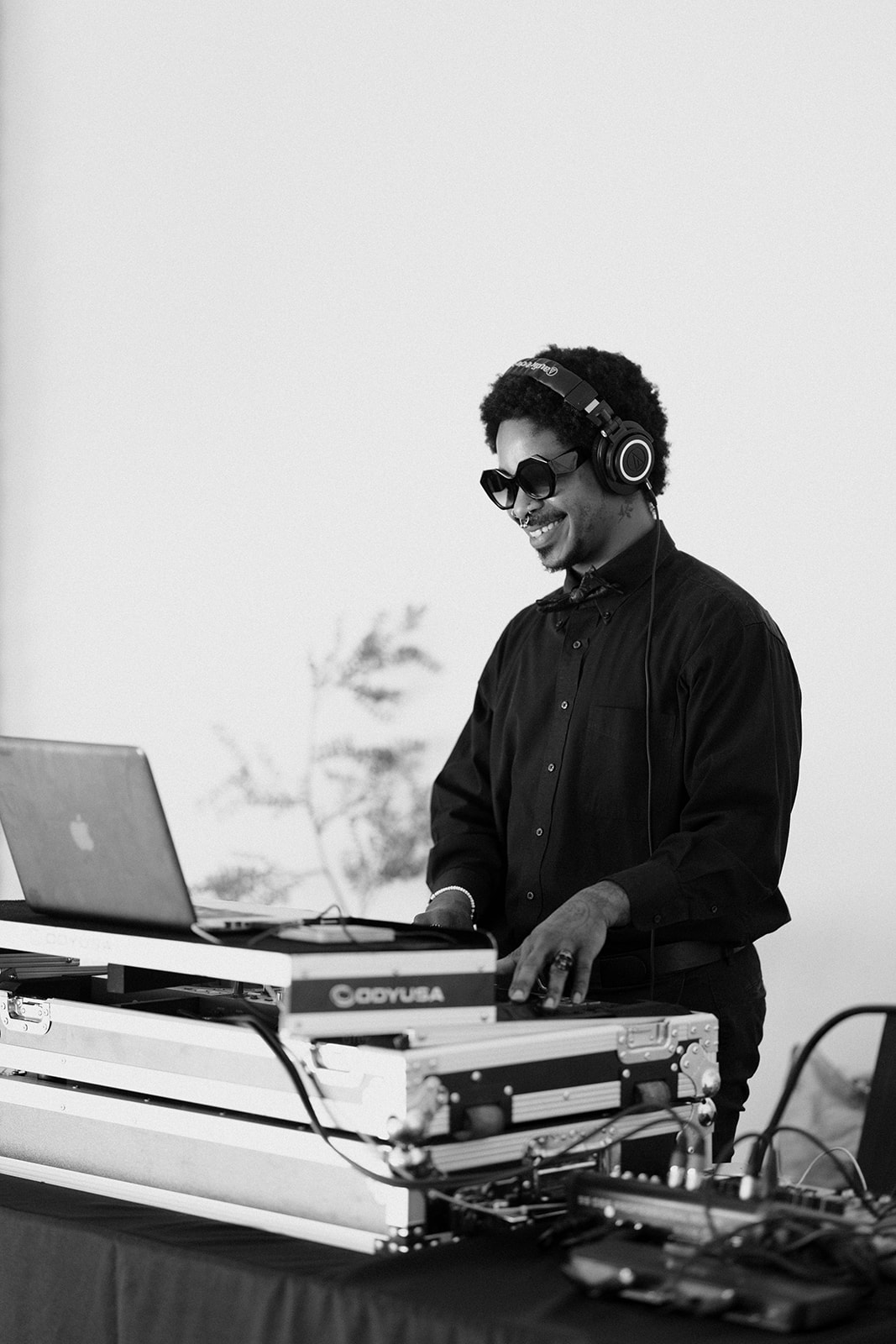 Person wearing headphones and sunglasses, smiling while operating DJ equipment, with a laptop and outdoor background.