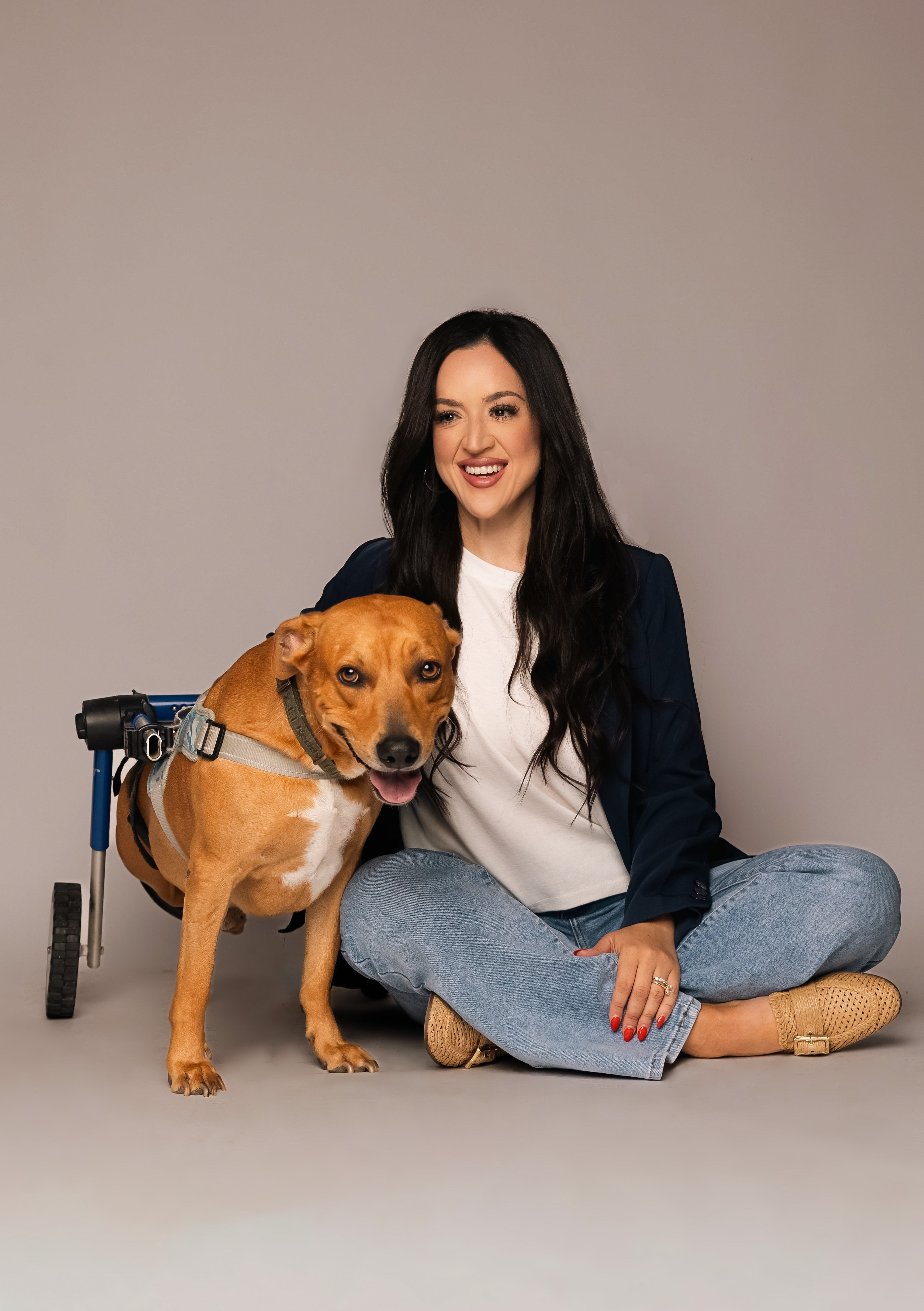 Woman sitting cross-legged on the floor with a brown dog wearing a harness, both smiling, against a plain background.