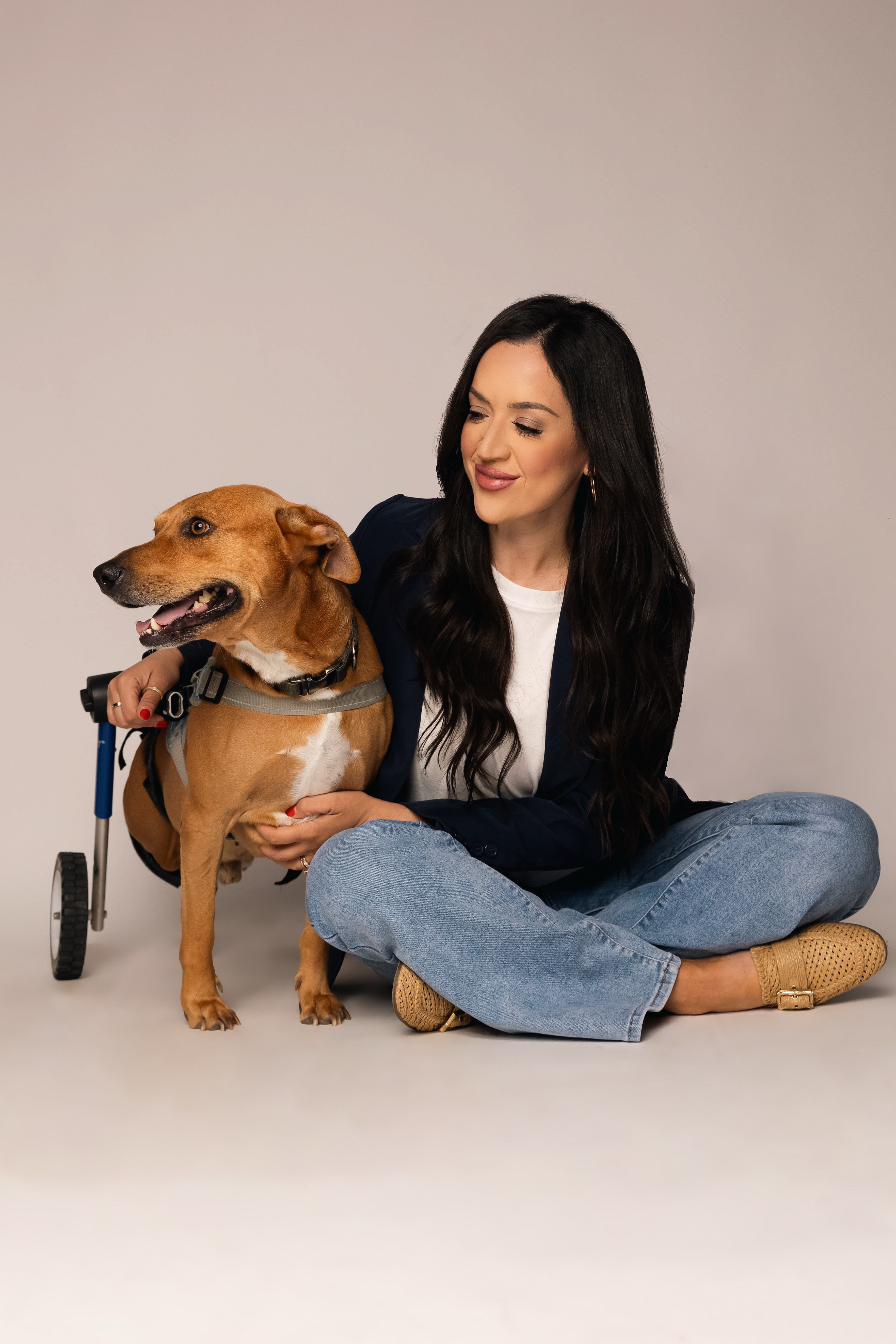 Woman sitting on the floor with a brown dog on a leash, both smiling, neutral background.