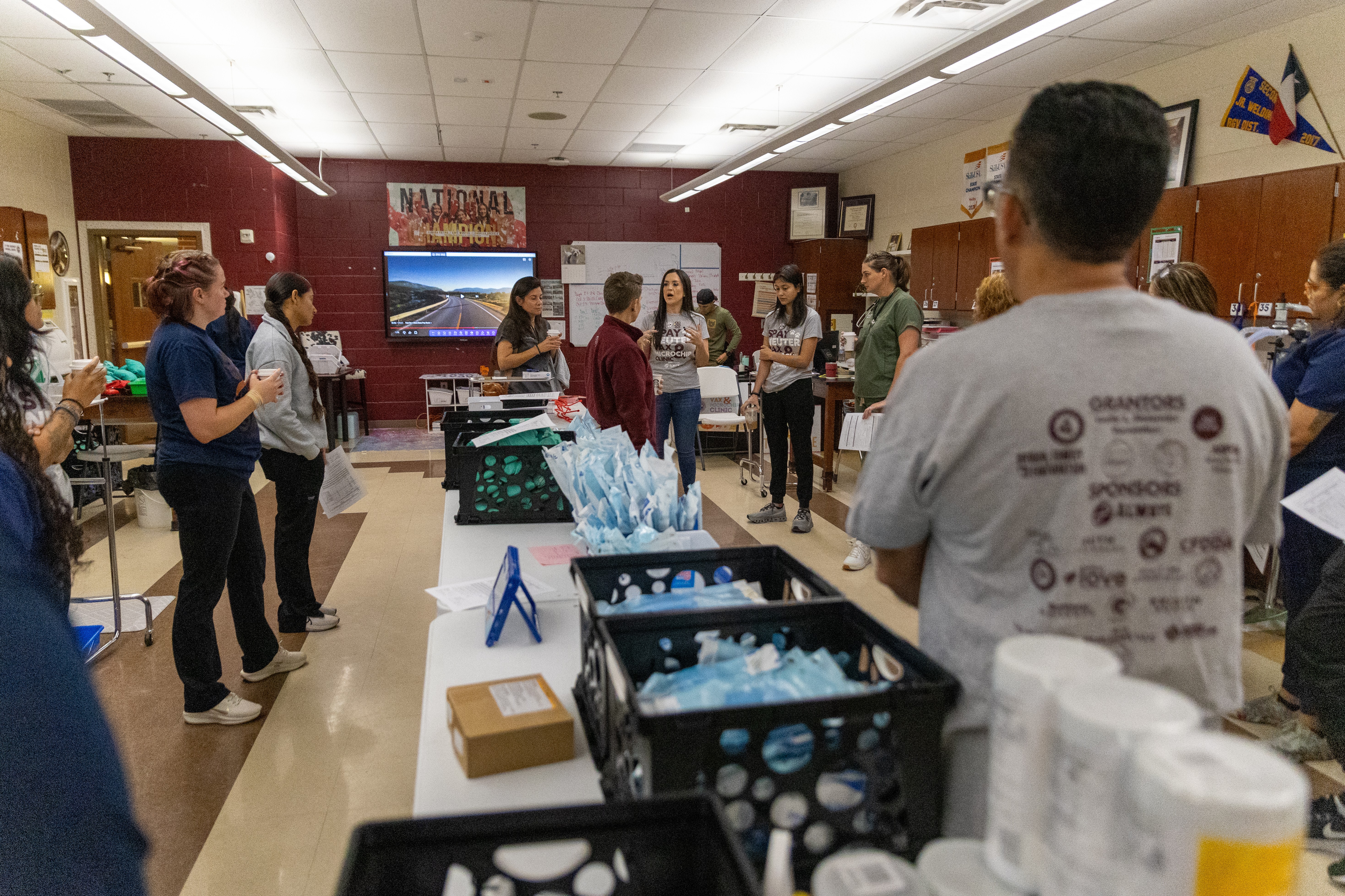 People gathered in a room with tables, water bottles, and a banner on the wall, engaging in an event or presentation.