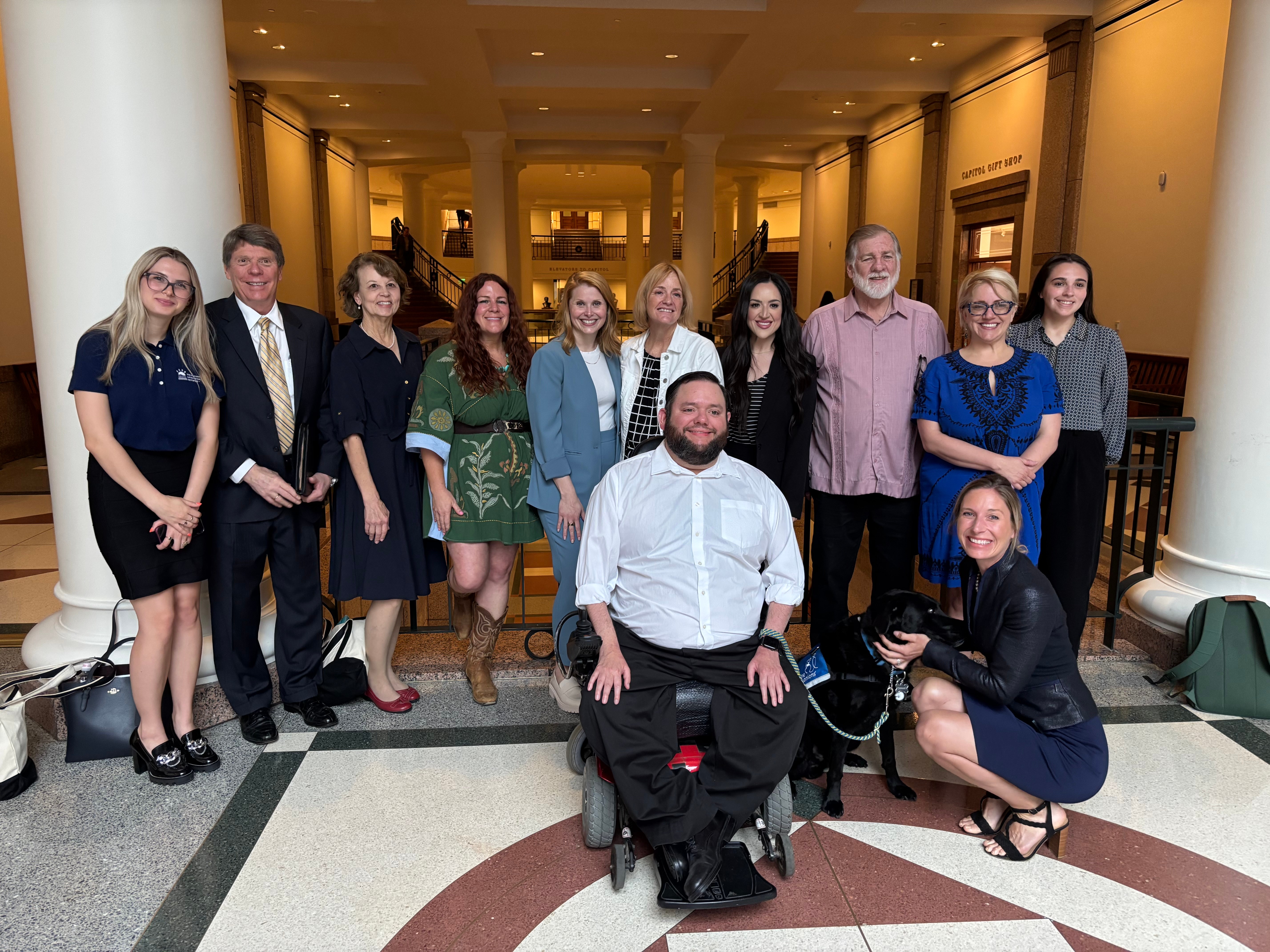 Group of ten people standing and sitting in a hallway, smiling, dressed in formal and casual attire, with a wheelchair user in the center.