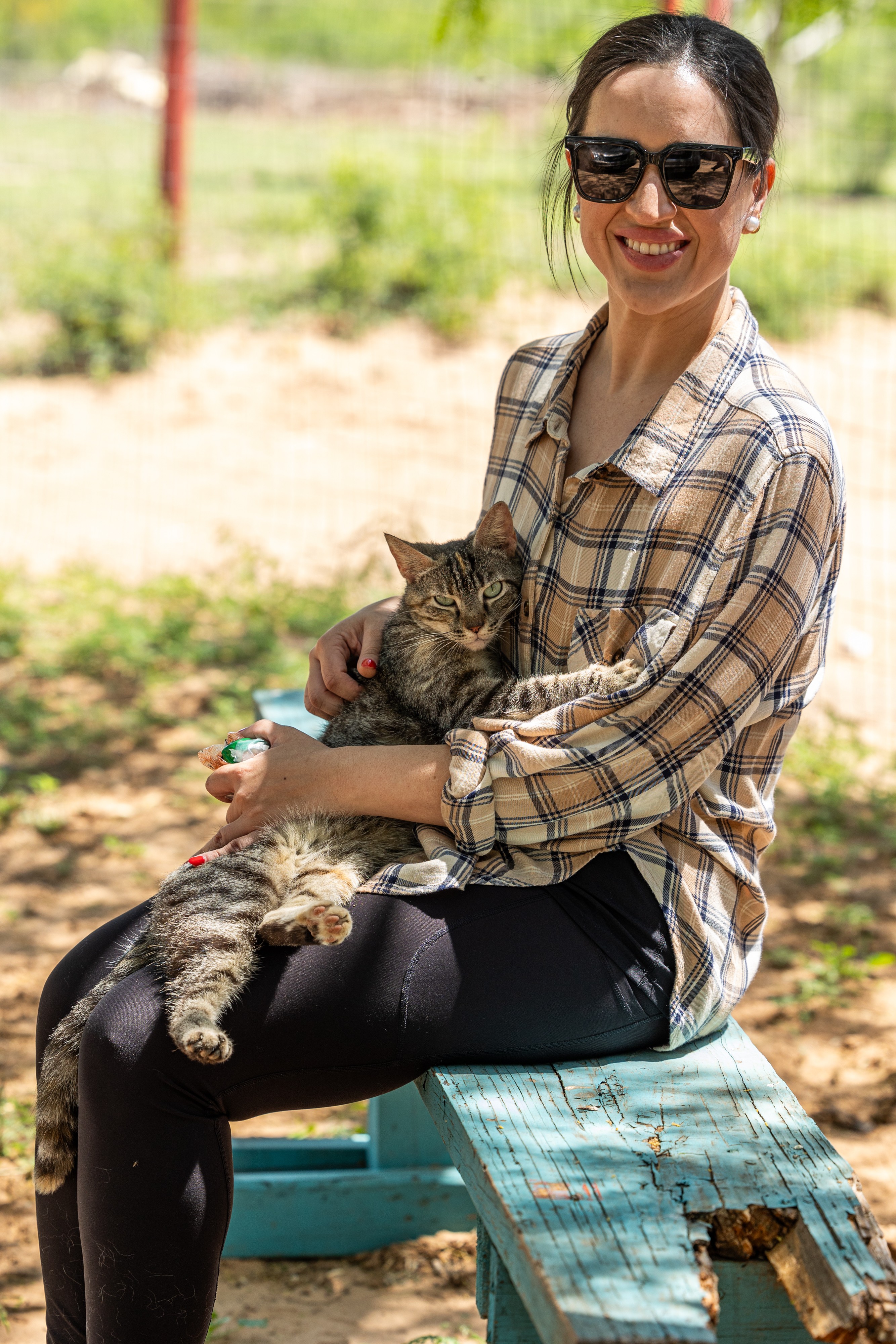 Woman sitting outdoors holding a tabby cat, smiling, wearing sunglasses and a plaid shirt.