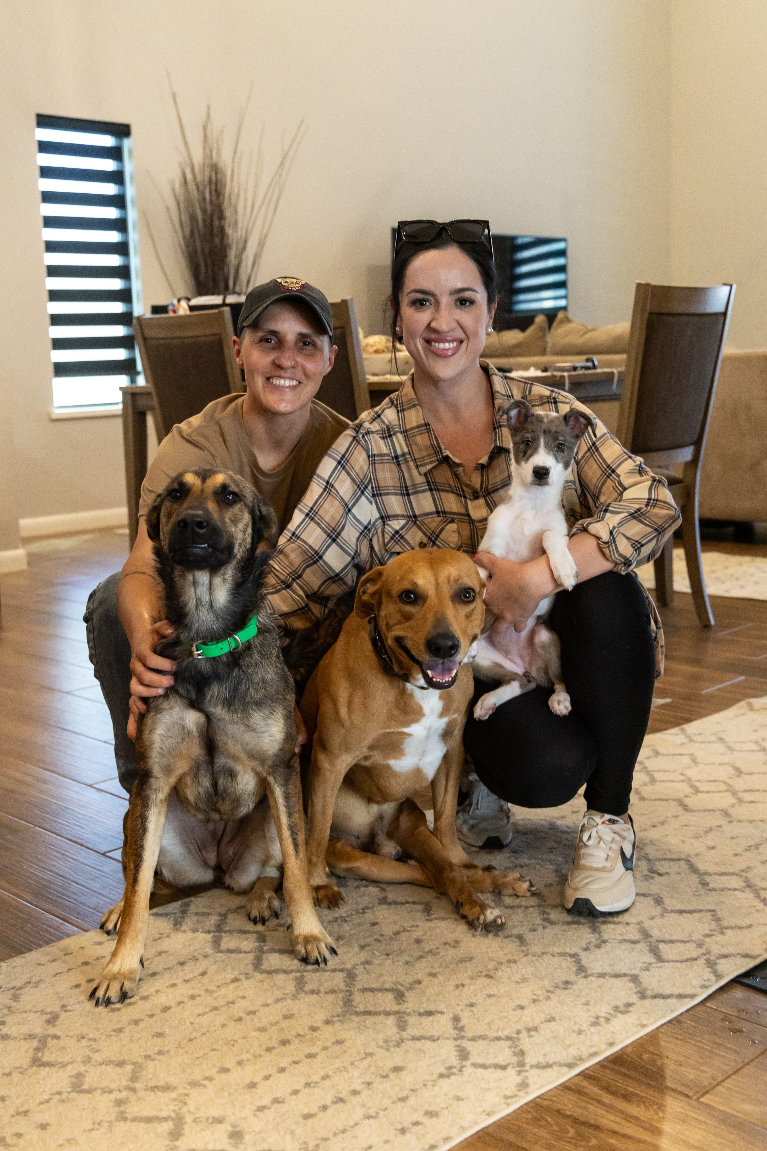 Two women with three dogs in a living room, smiling, with a dining table and chairs in the background.