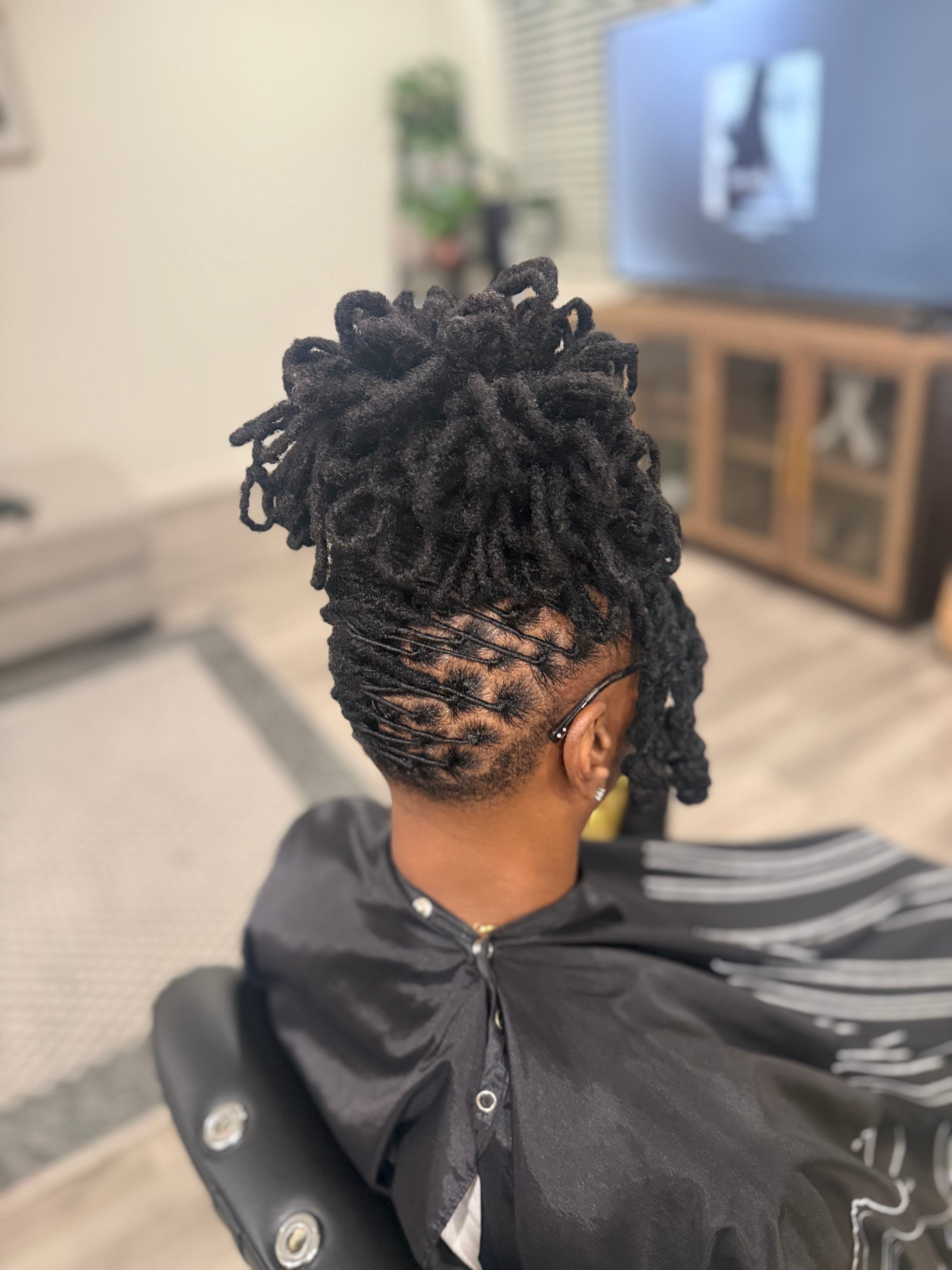 Person with styled dreadlocks sitting in a salon chair, facing away, with a television and furniture in the background.