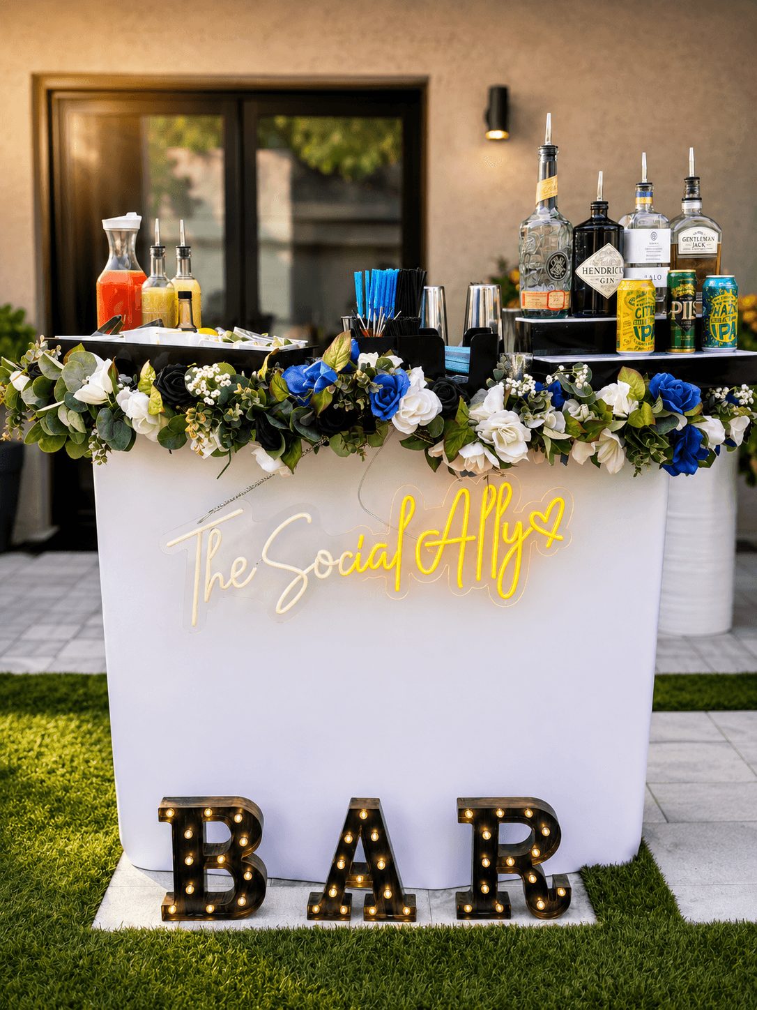 Outdoor bar setup with floral decorations, bottles, and a neon sign reading 'The Social Ally'. Large illuminated 'BAR' letters on grass.