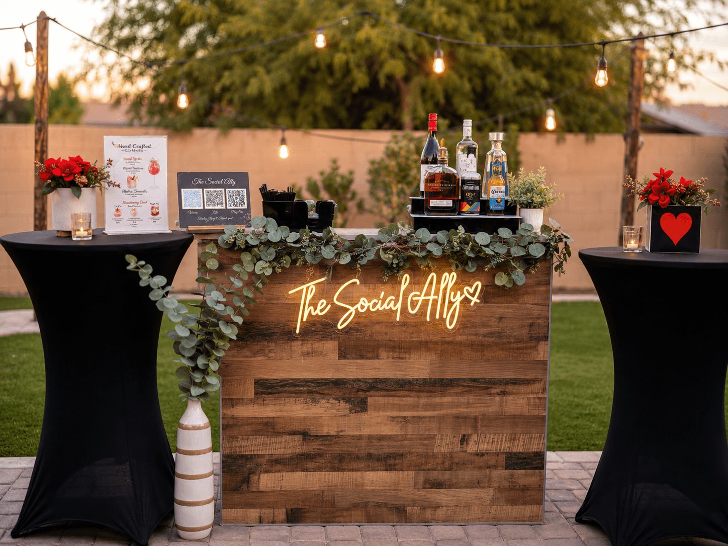 Outdoor bar setup with a wooden counter, string lights, and two tall tables with black covers and red flower arrangements.