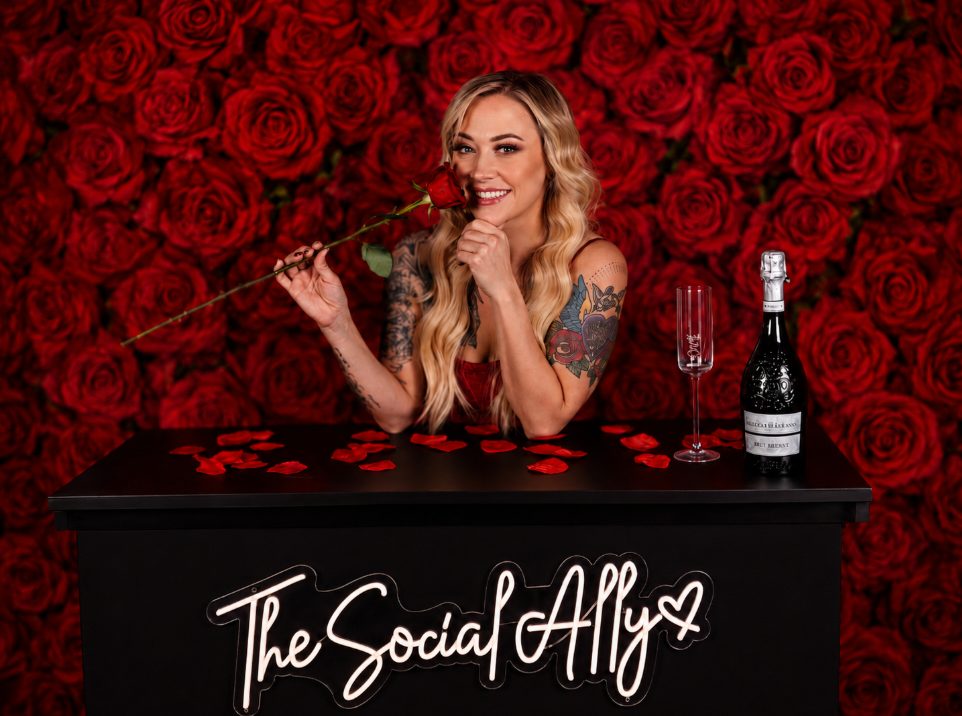 Woman with long blonde hair holding a rose, smiling, with a red rose background, a champagne bottle, and a glass on a black table.