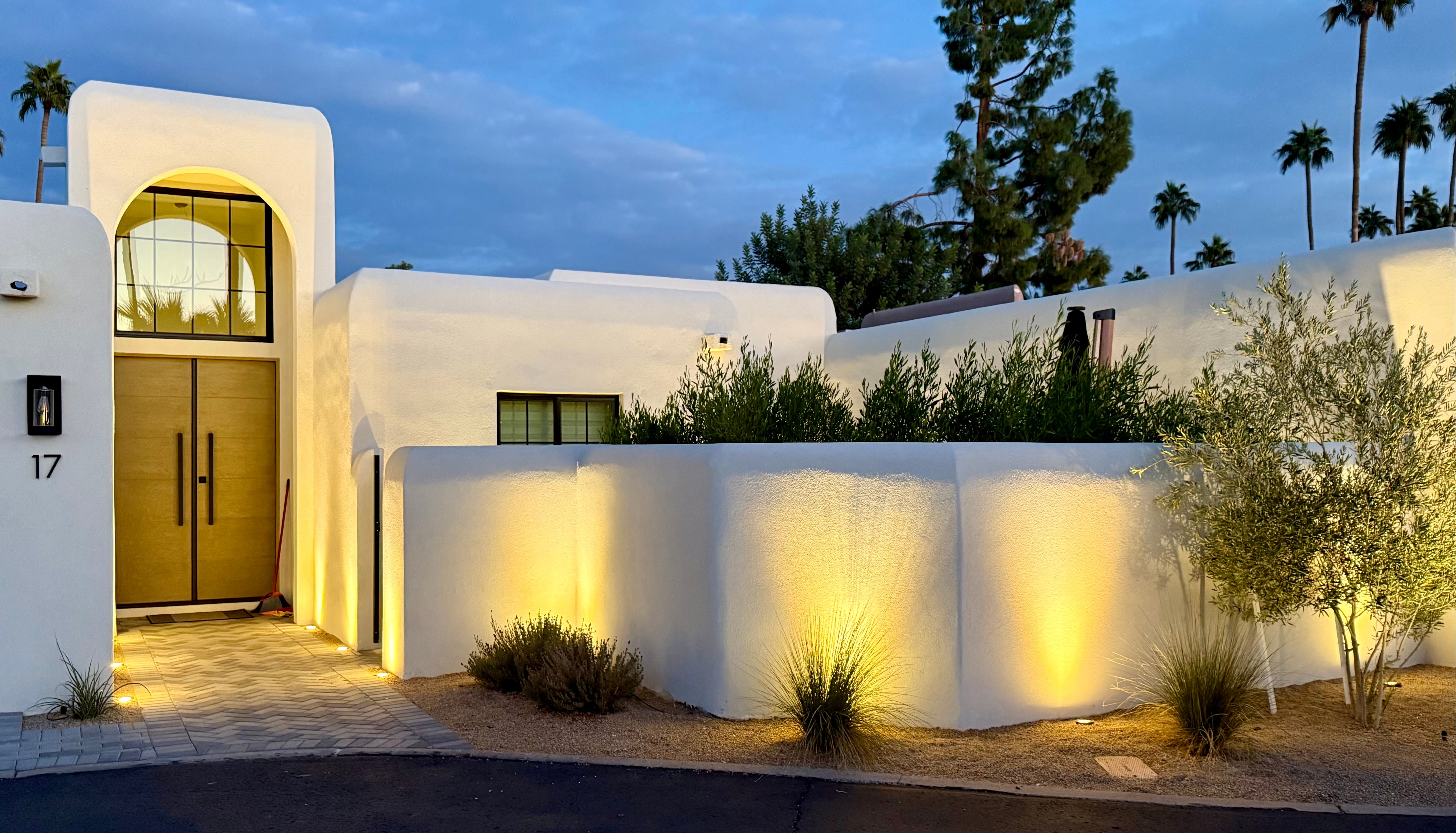 Modern white building with curved walls, illuminated plants, and a wooden door, under a blue sky with palm trees.