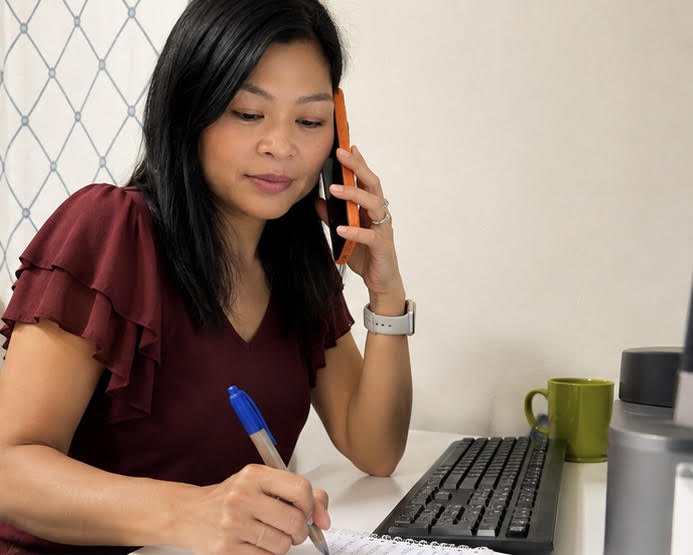 Woman with black hair, wearing a maroon top, talking on a phone, writing with a pen, sitting at a desk with a keyboard and mug.