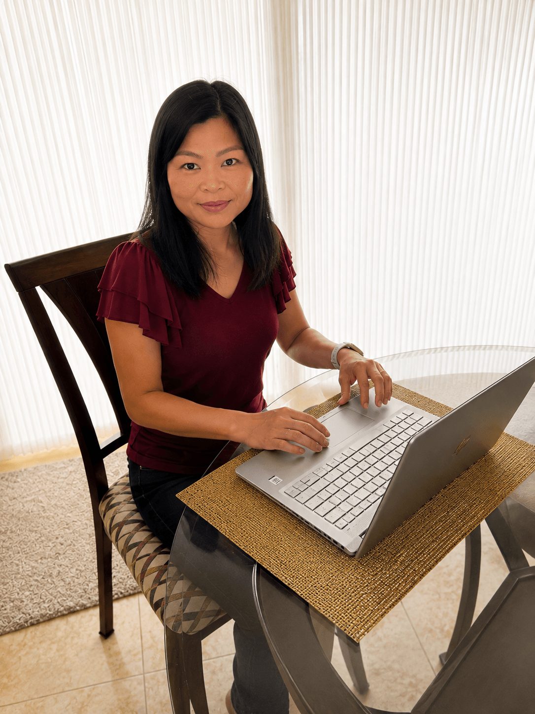 Woman sitting at a table with a laptop, smiling, in a room with vertical blinds.