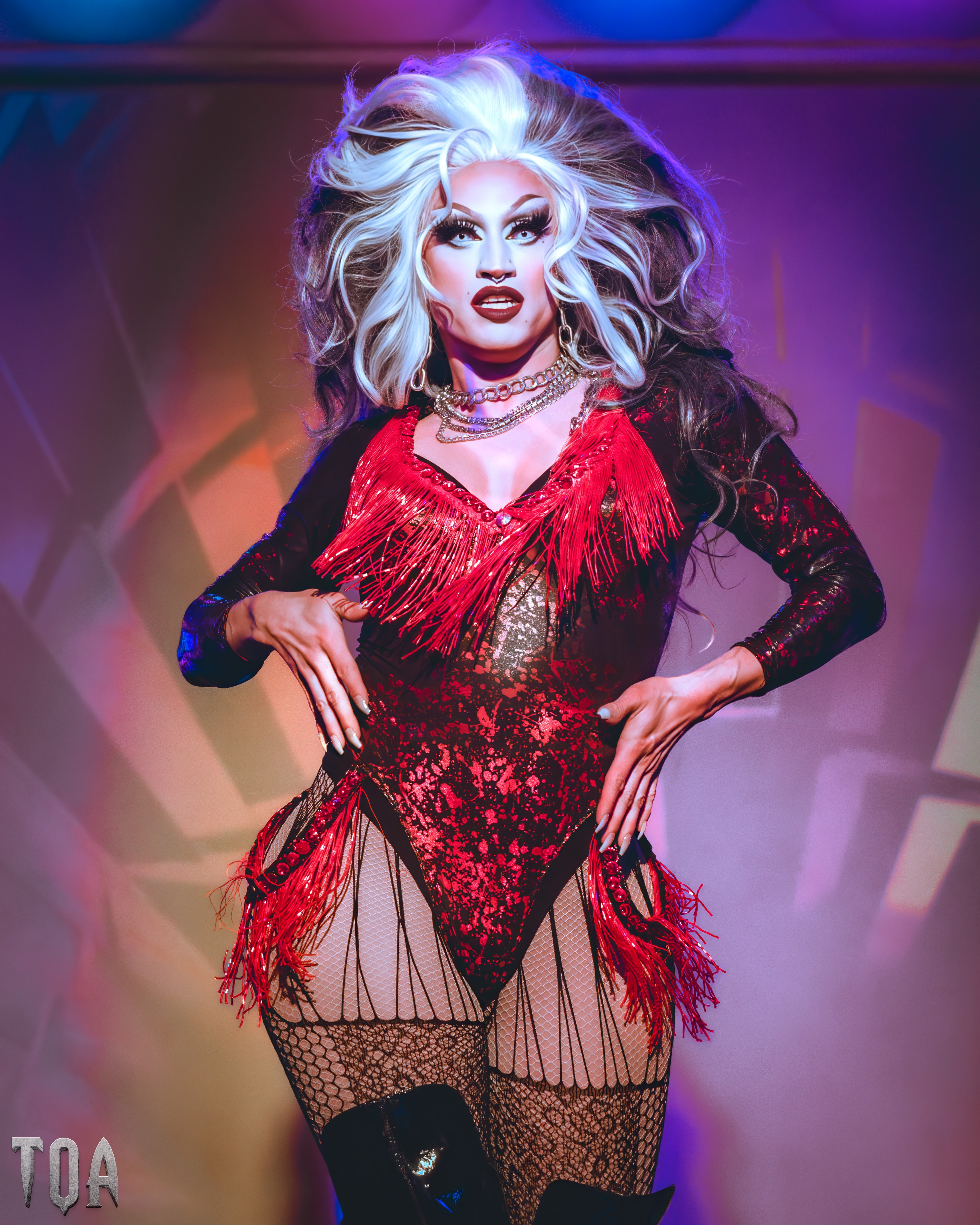 Person with voluminous white and black hair, wearing a red and black lace outfit with fishnet stockings, posing confidently.