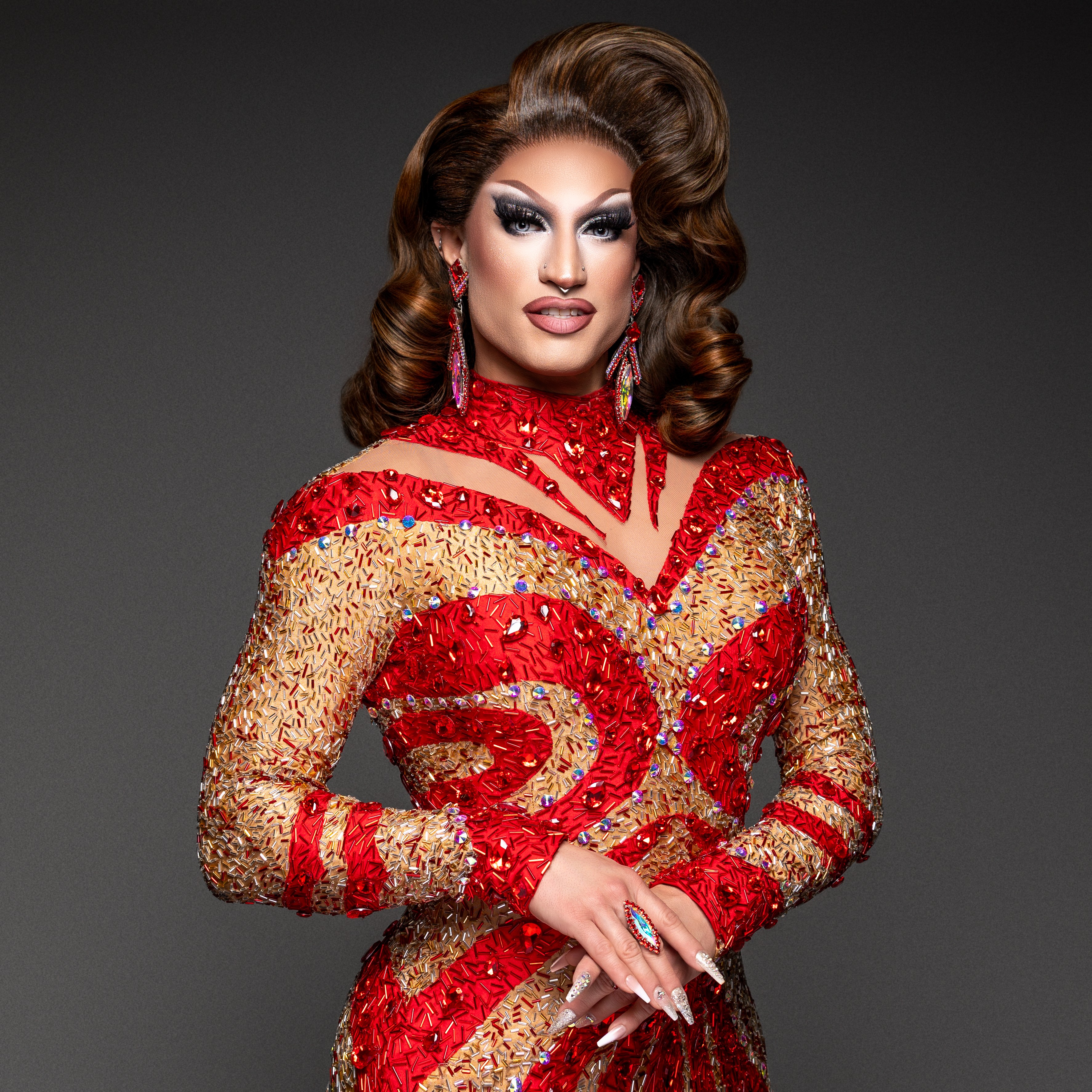 A person with styled hair and makeup wearing a red and gold embellished dress with long sleeves and earrings.