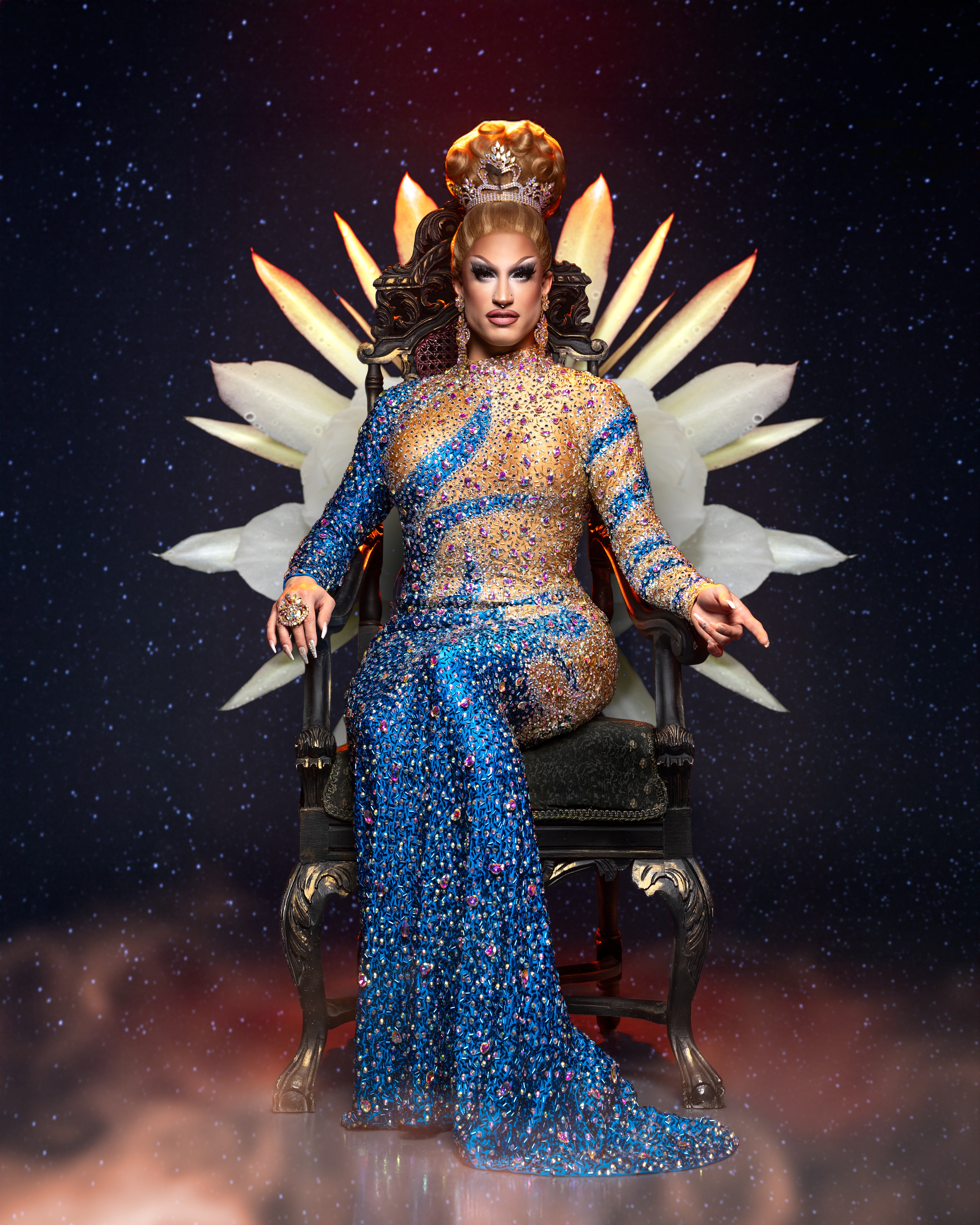 Person with elaborate hairstyle and makeup sitting on a throne with a starry background, wearing a blue and gold gown.