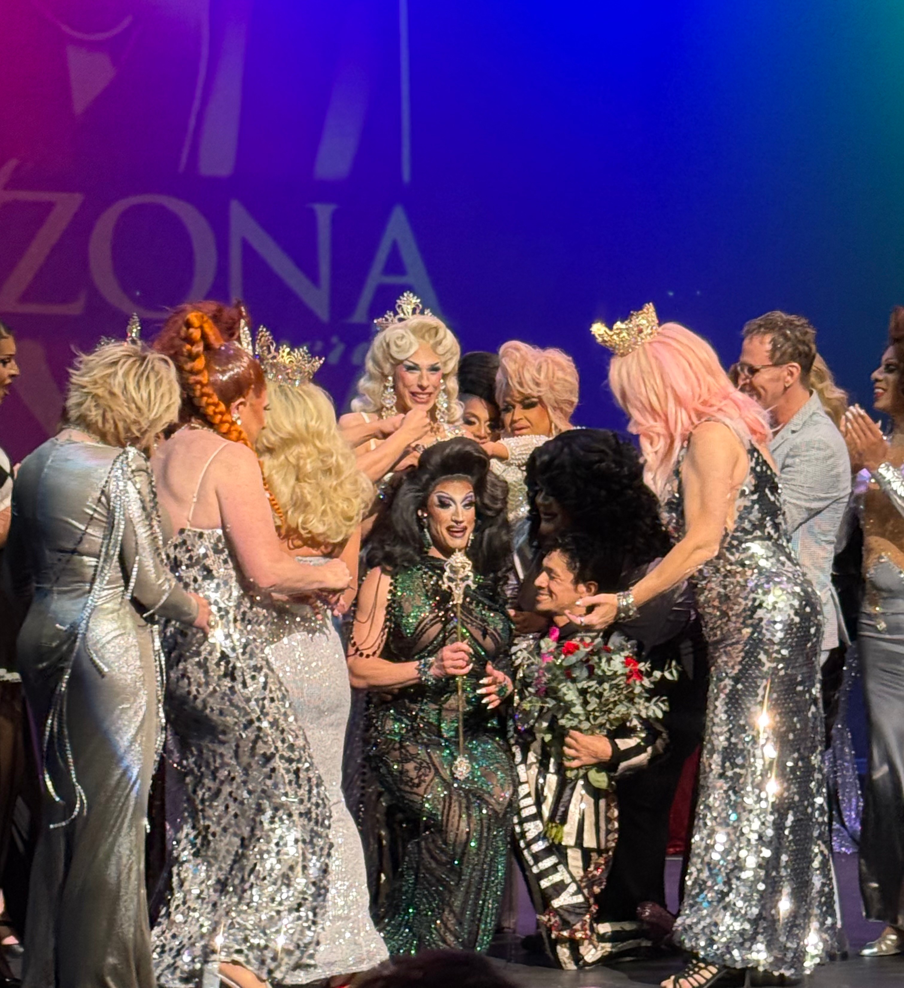 Group of people dressed in glamorous, shiny outfits, some wearing crowns, gathered on stage with a purple backdrop.