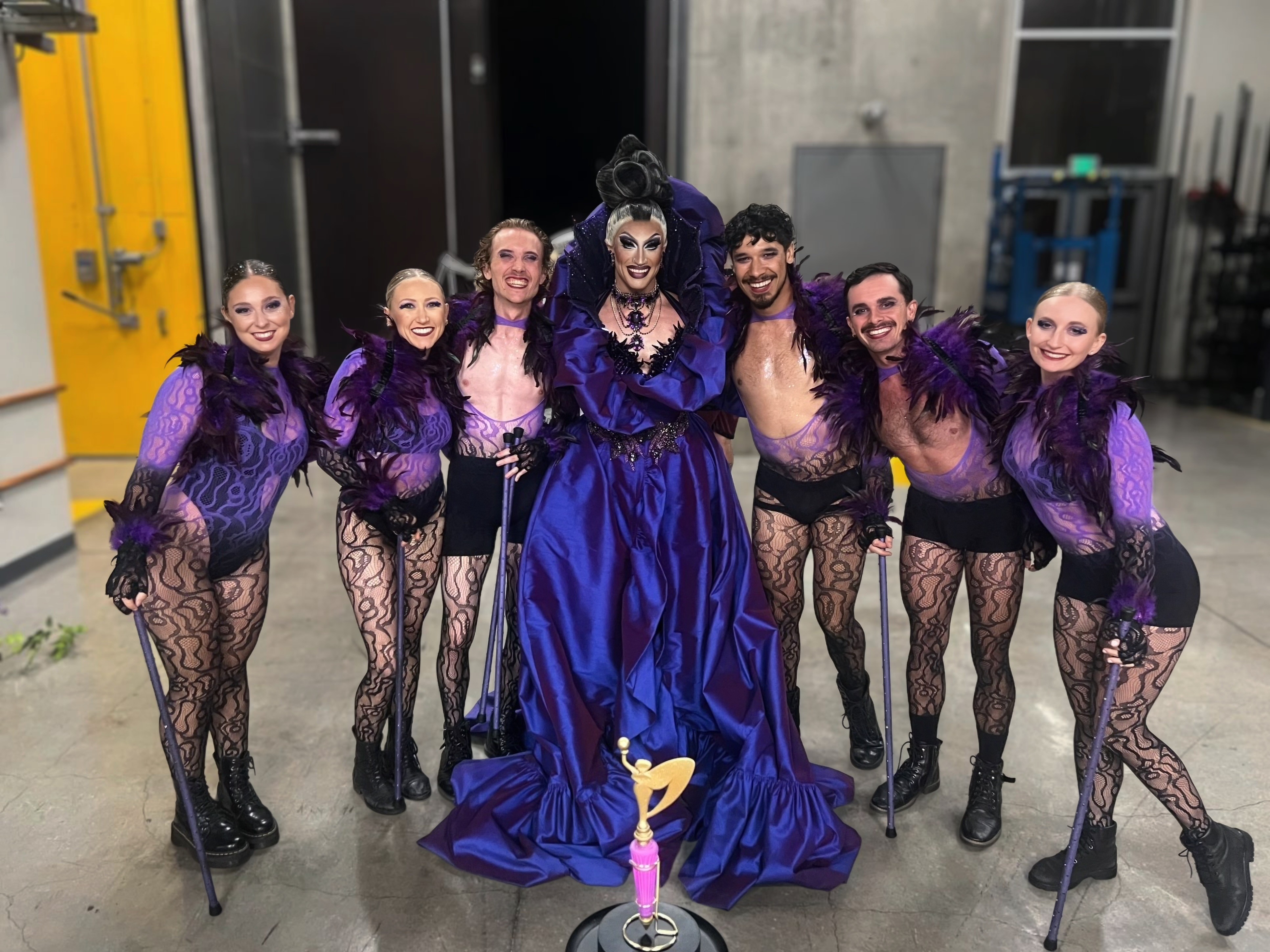 Group of seven performers in costumes, standing together backstage, with a woman in a large blue gown in the center.