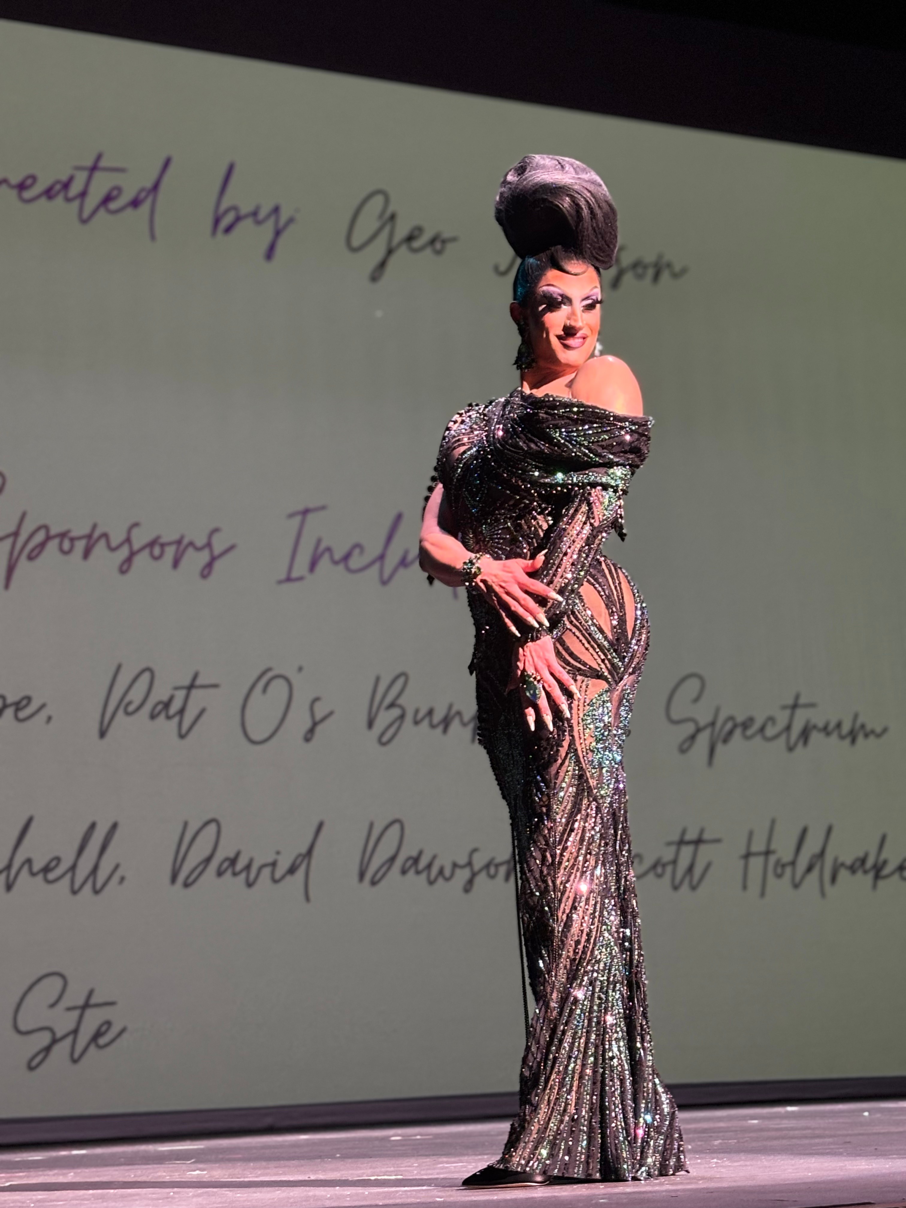 Person in a shiny, patterned gown with an elaborate hairstyle standing on stage in front of a screen with handwritten text.