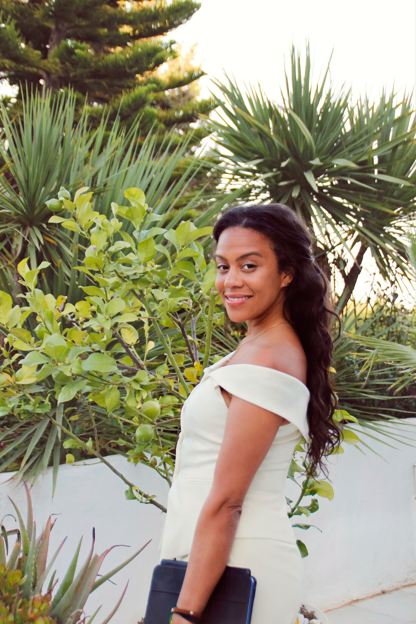 Young woman with long dark hair in a white off-shoulder dress standing outdoors among green plants and trees, smiling.