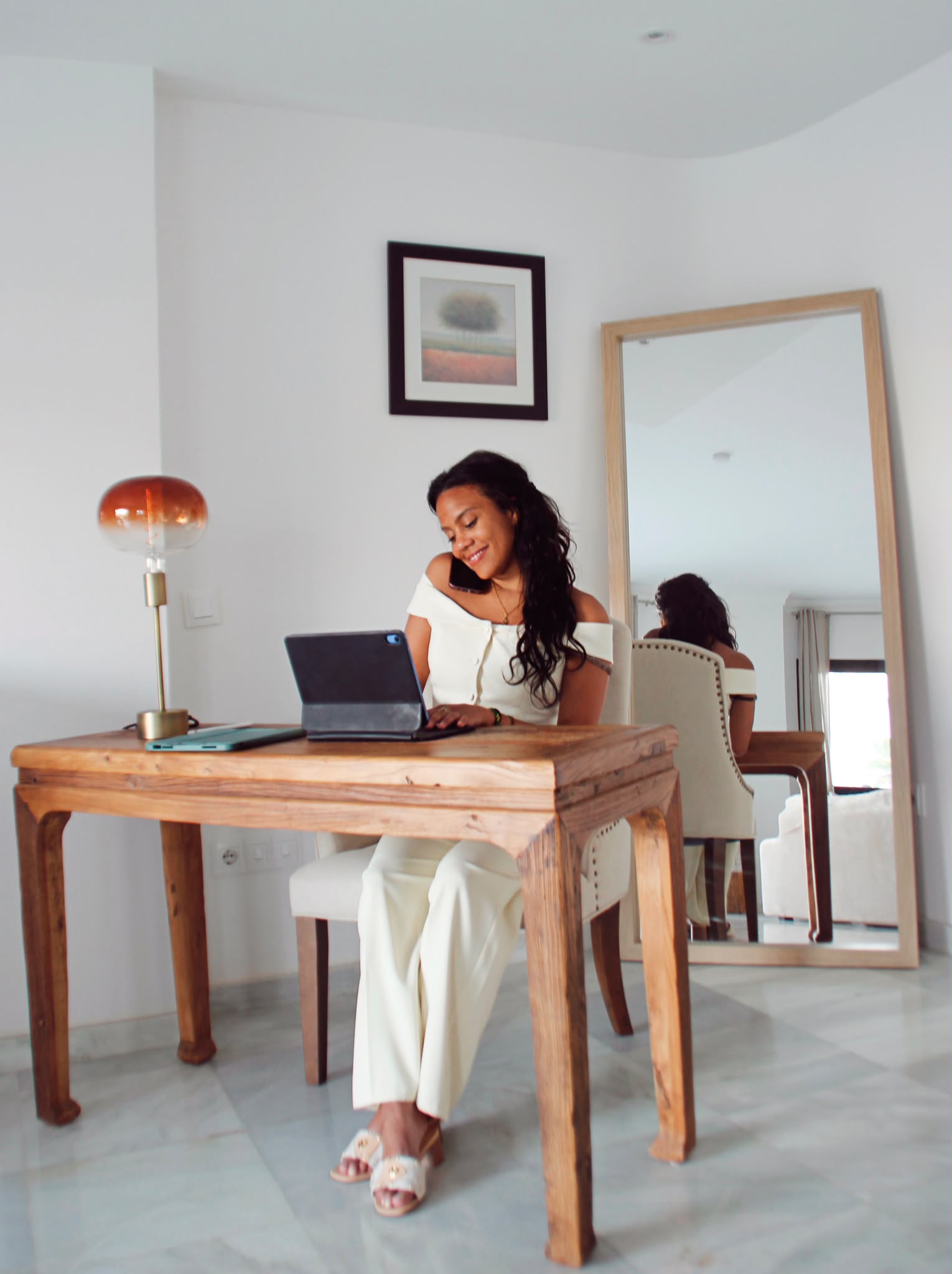 Woman sitting at a wooden table using a laptop, with a mirror and framed picture on the wall behind her.