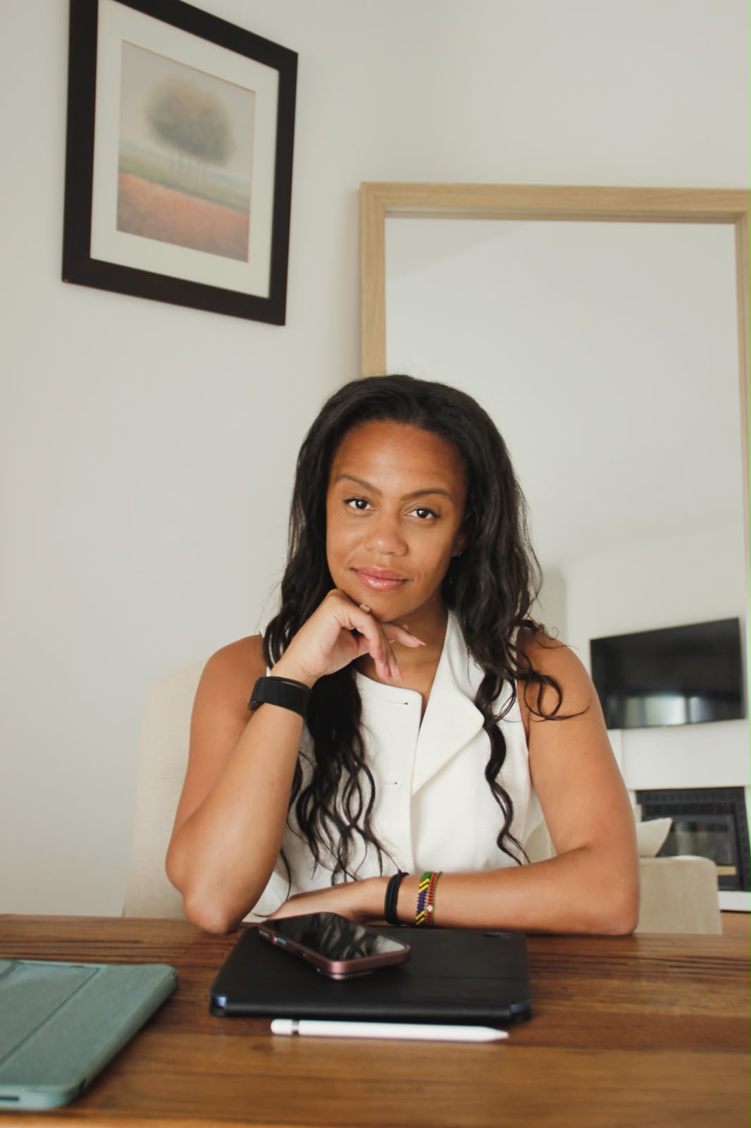 Young woman with long dark hair sitting at a table, resting her chin on her hand, in a room with framed artwork on the wall.