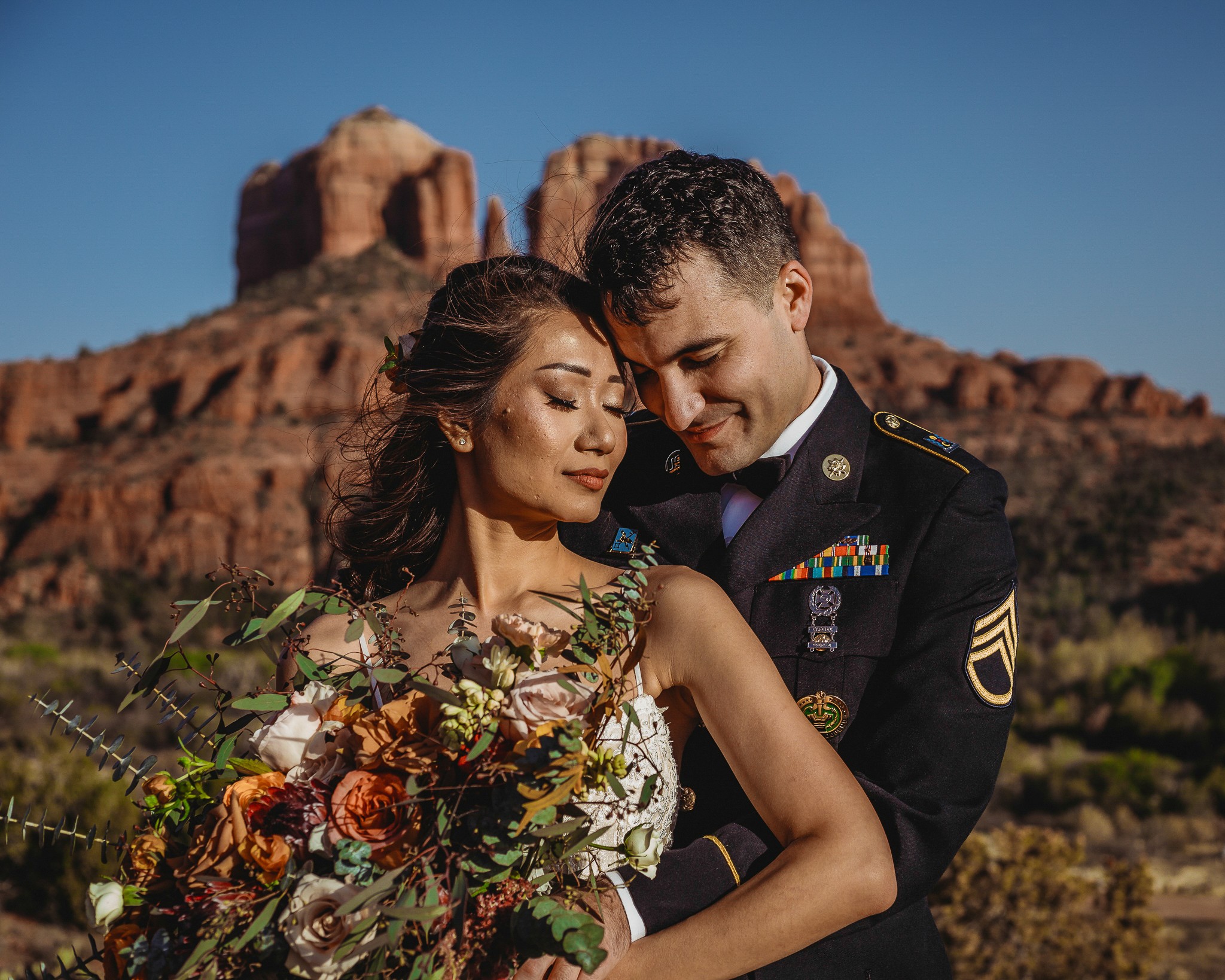A couple, one in military uniform, embraces outdoors with a rocky mountain in the background.